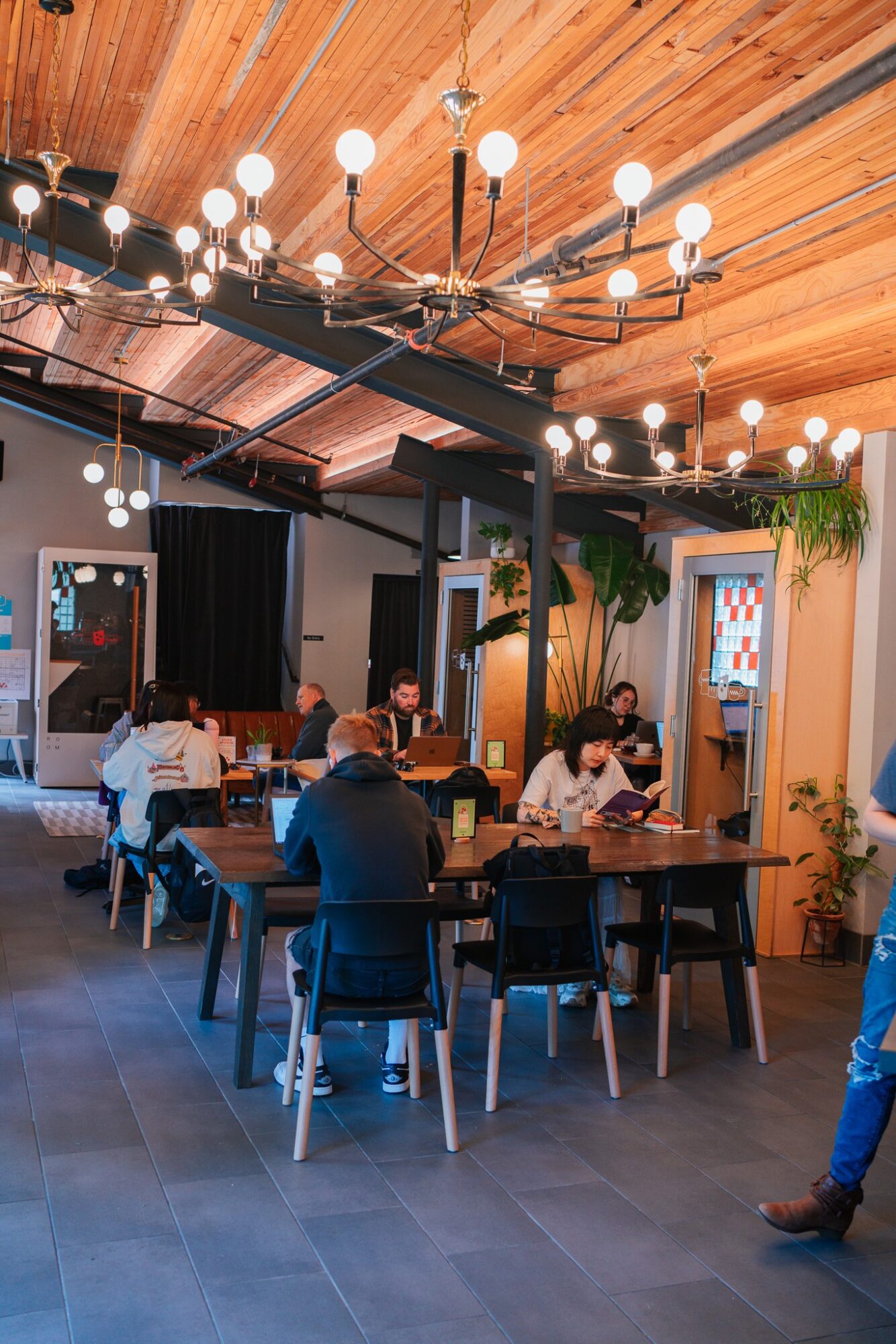 People sit at tables in a modern indoor space with wooden ceiling, large chandelier, and decorative plants.