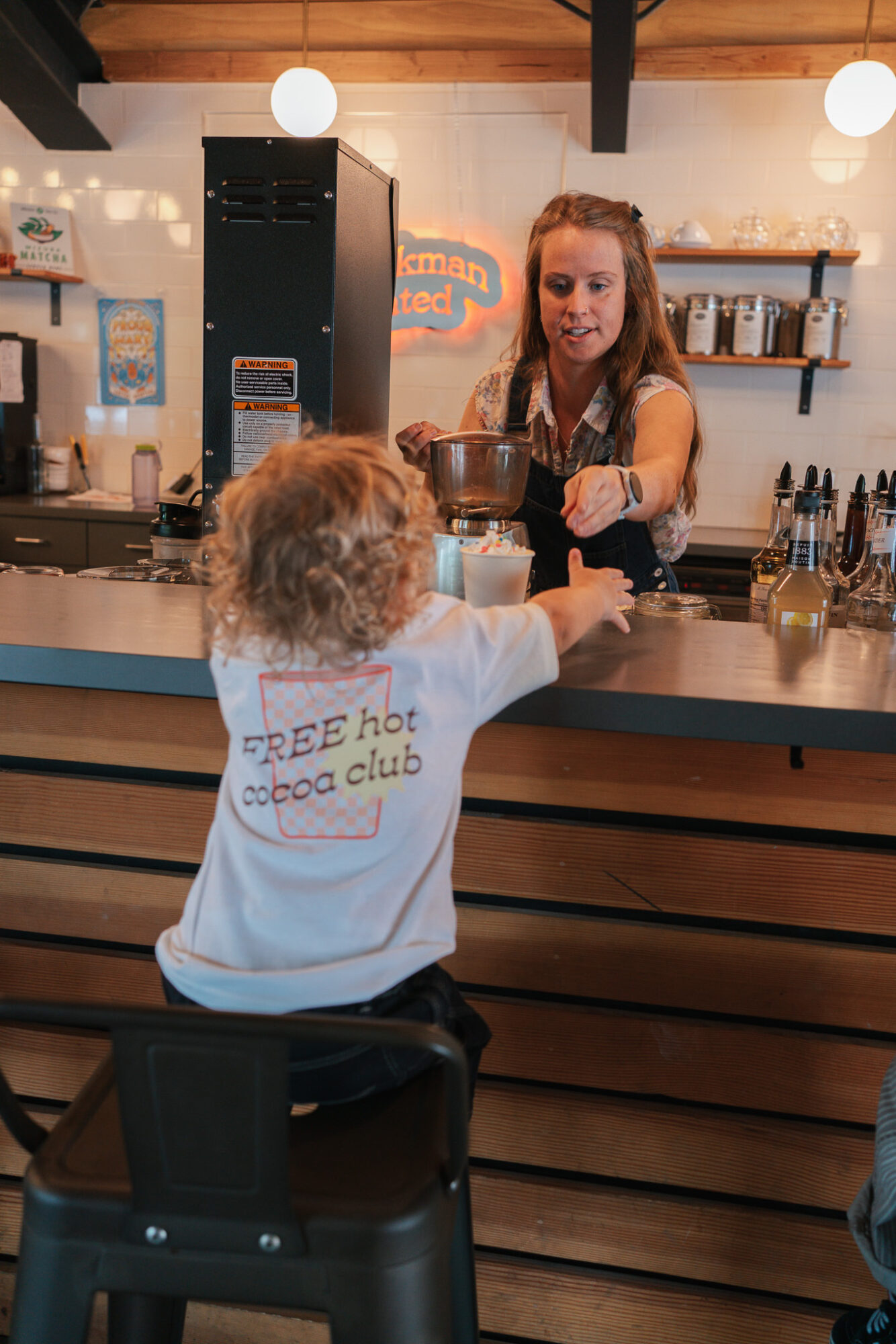 Child with curly hair and a white shirt at a counter, talking to a barista in a coffee shop.