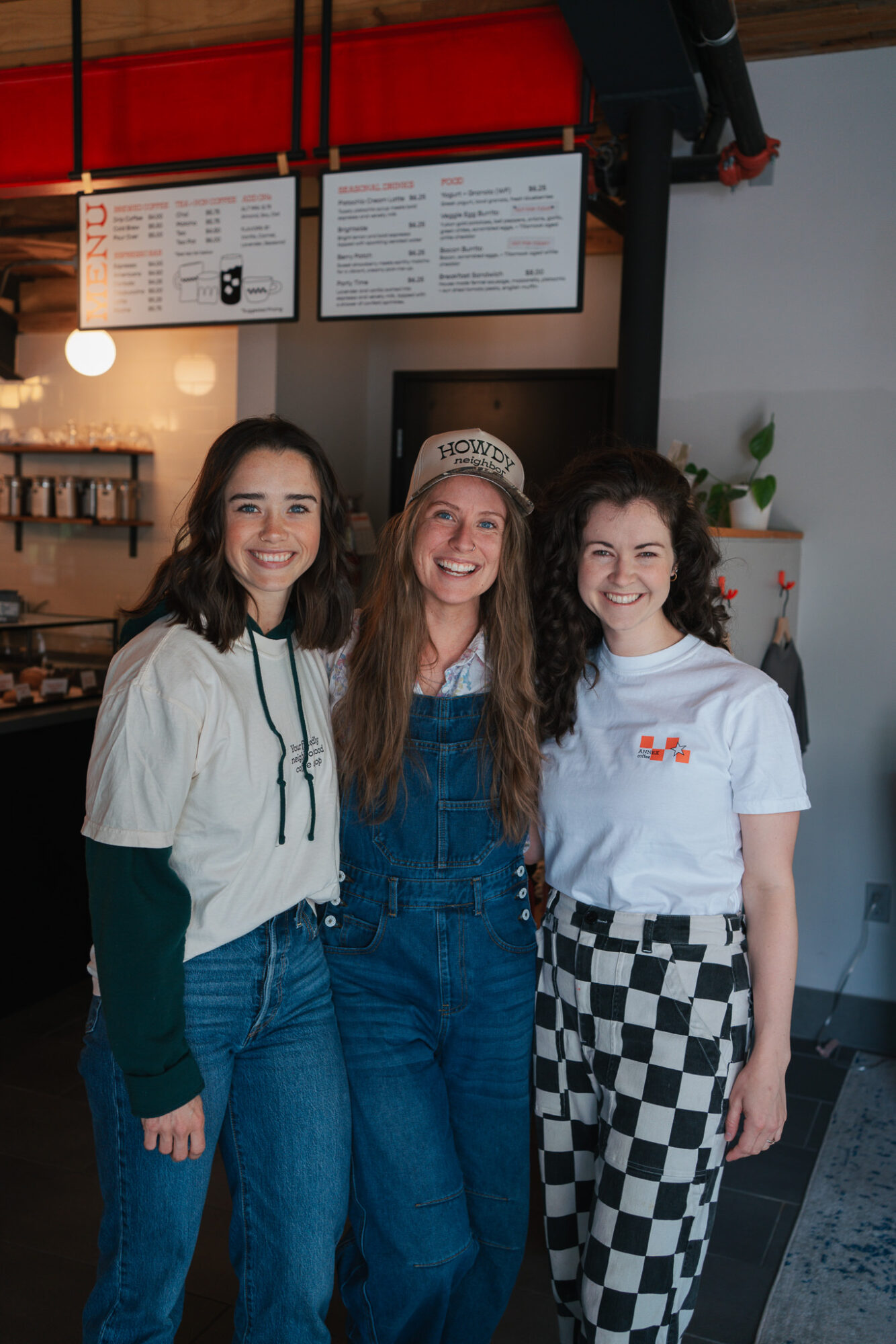 Three women smiling inside a cafe, standing close together, with menu boards above them and a plant in the background.
