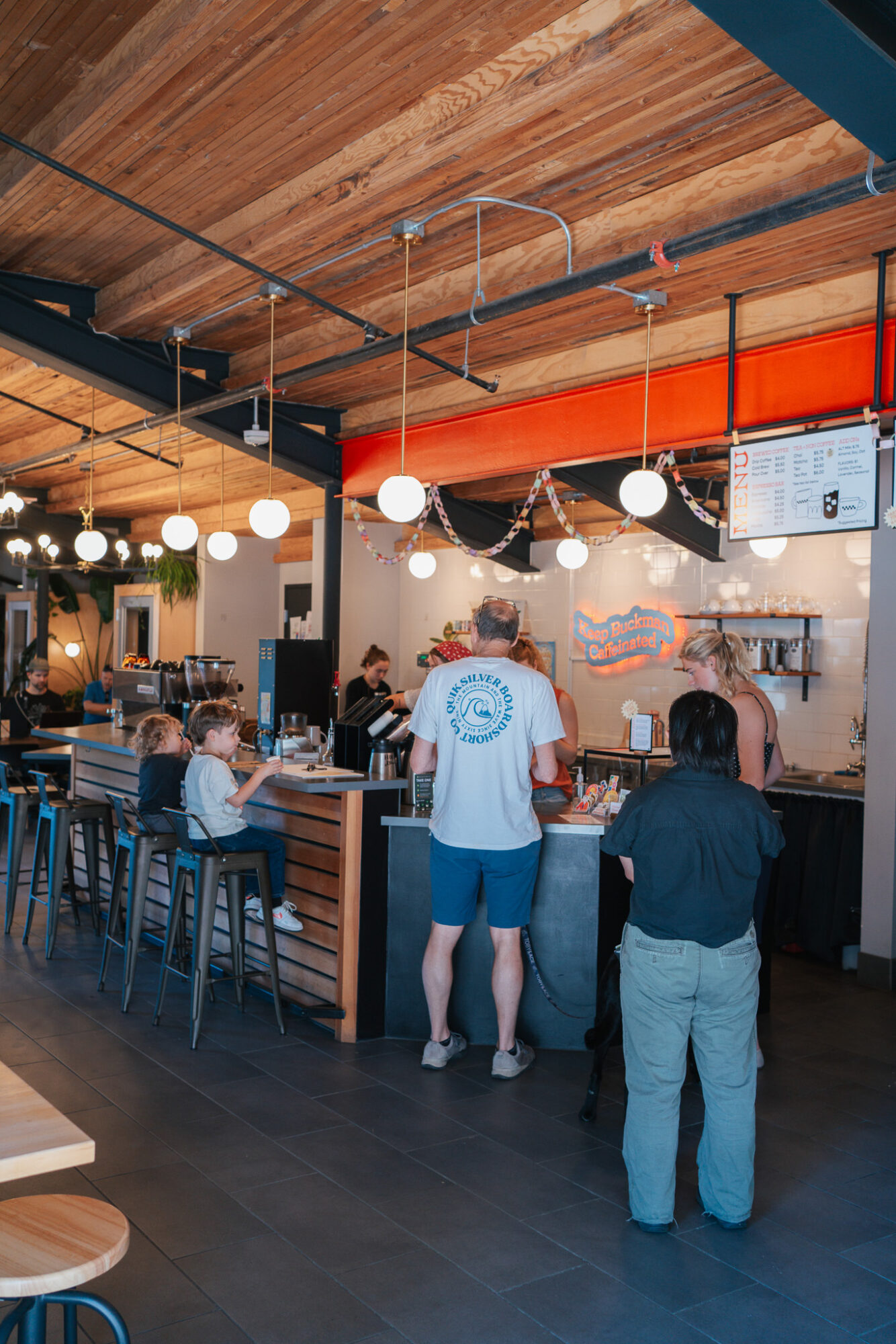 People sitting and standing at a bar counter in a modern restaurant with wooden ceiling and hanging lights.