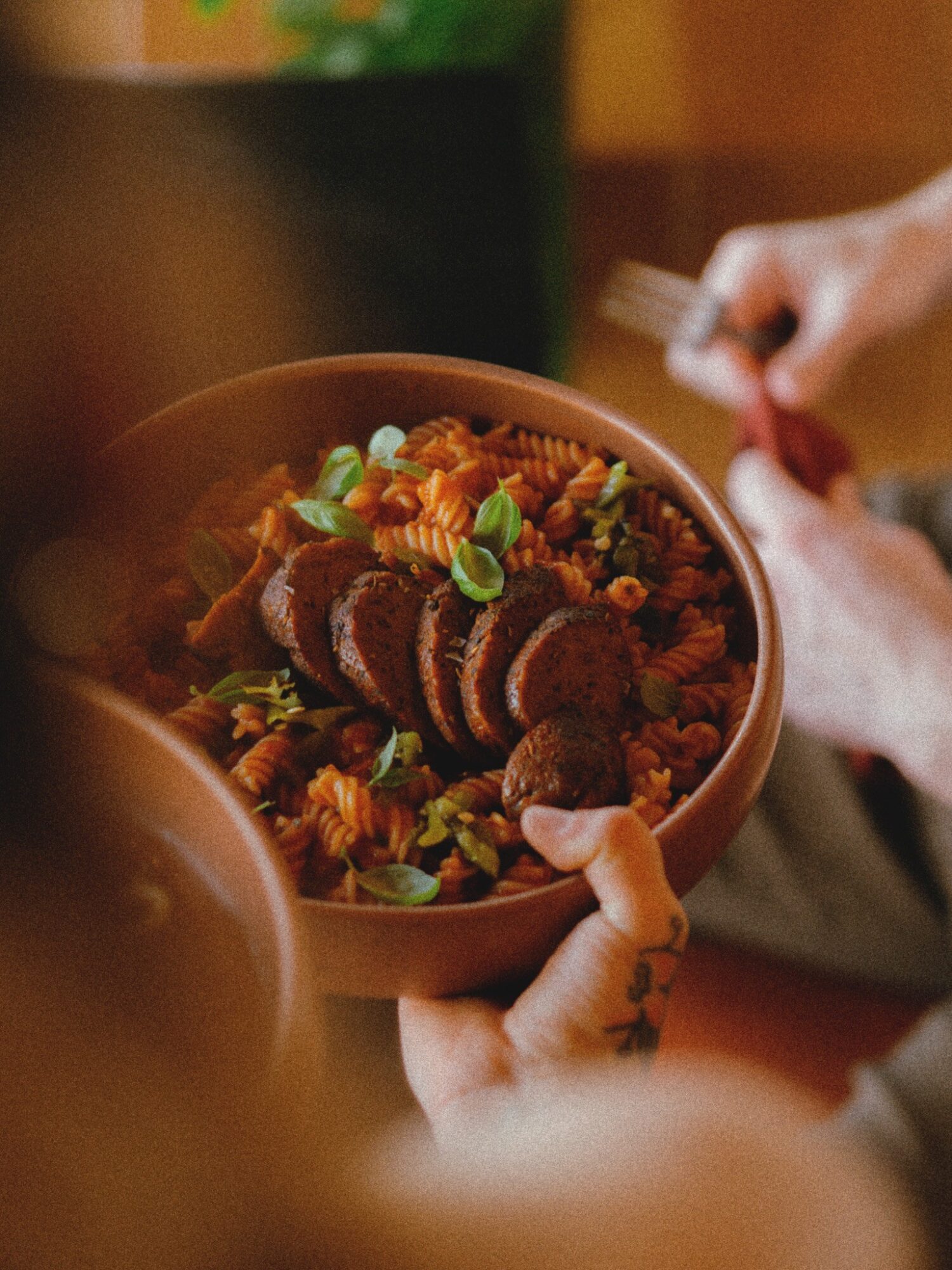 Person holding a bowl of pasta with sliced meat and green onions, with another person in the background holding a fork.