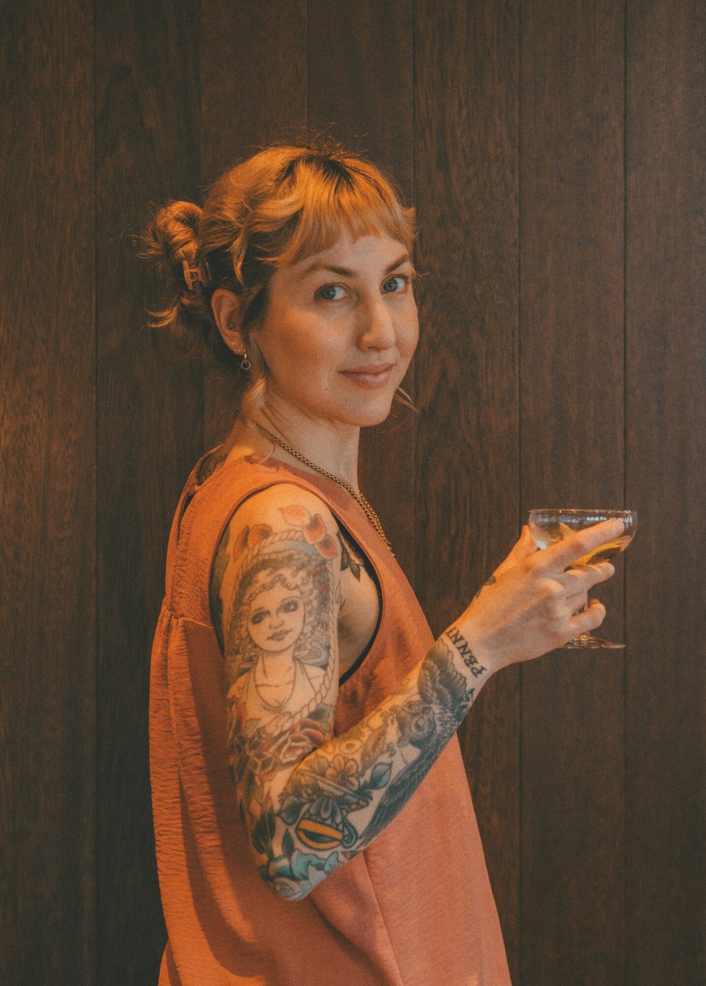Woman with tattoos on her arm holding a glass, standing against a dark wooden background.
