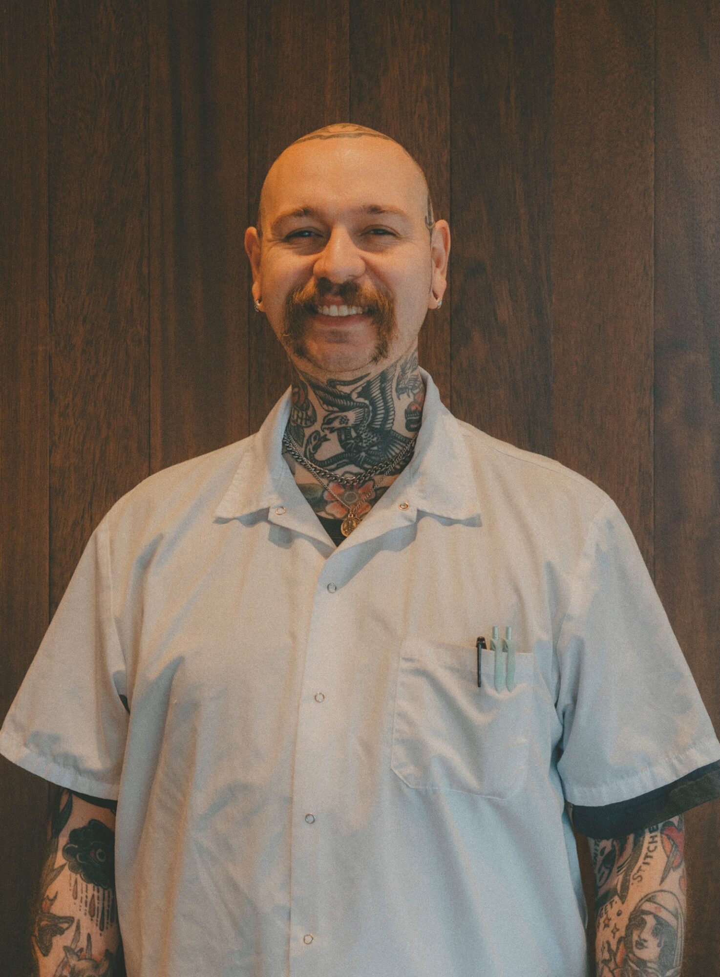 Man with tattoos on arms and neck, smiling, wearing a white shirt, standing against a wooden background.