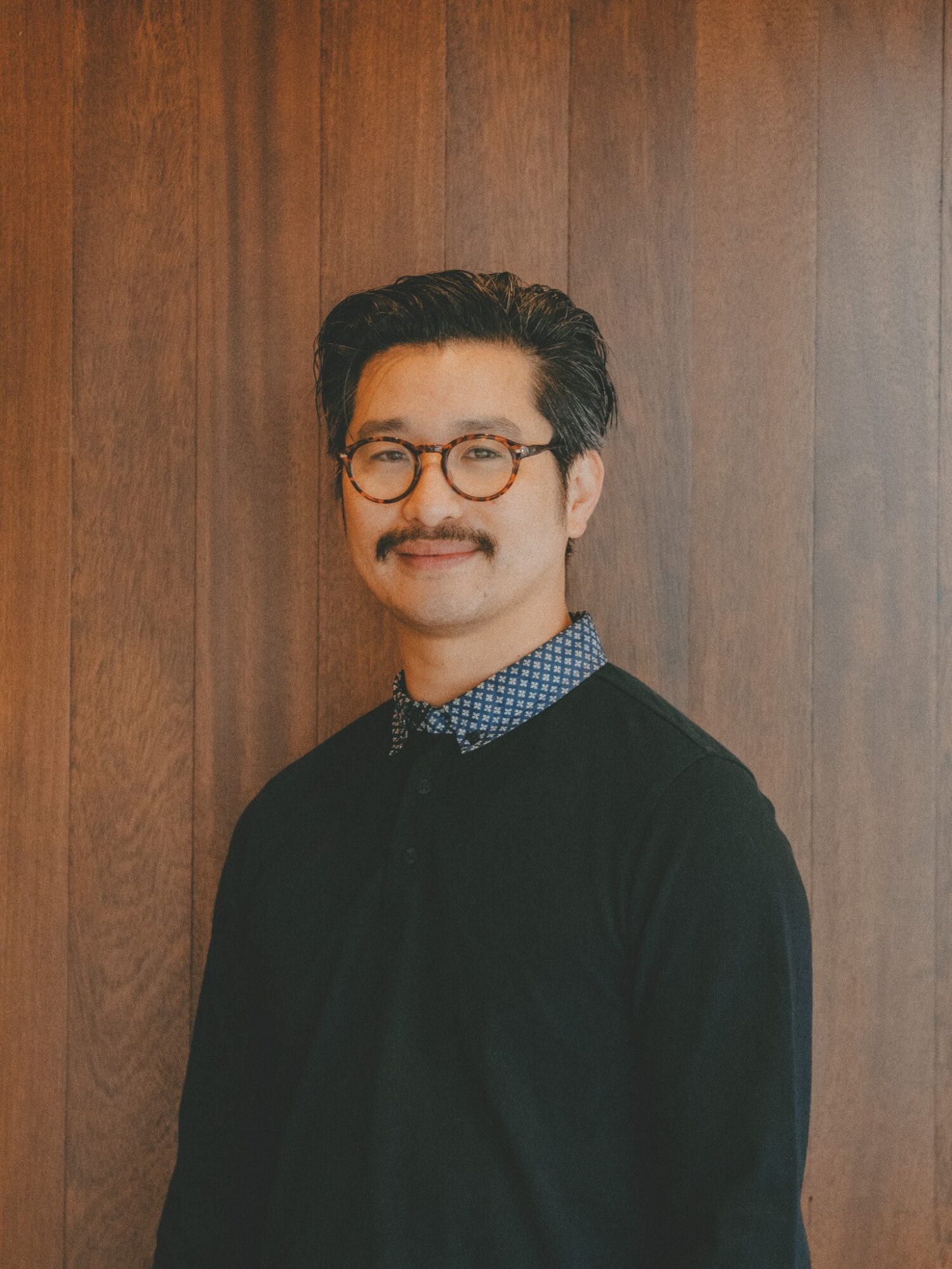 Man with glasses and a mustache, wearing a dark sweater over a patterned shirt, standing against a wooden wall.
