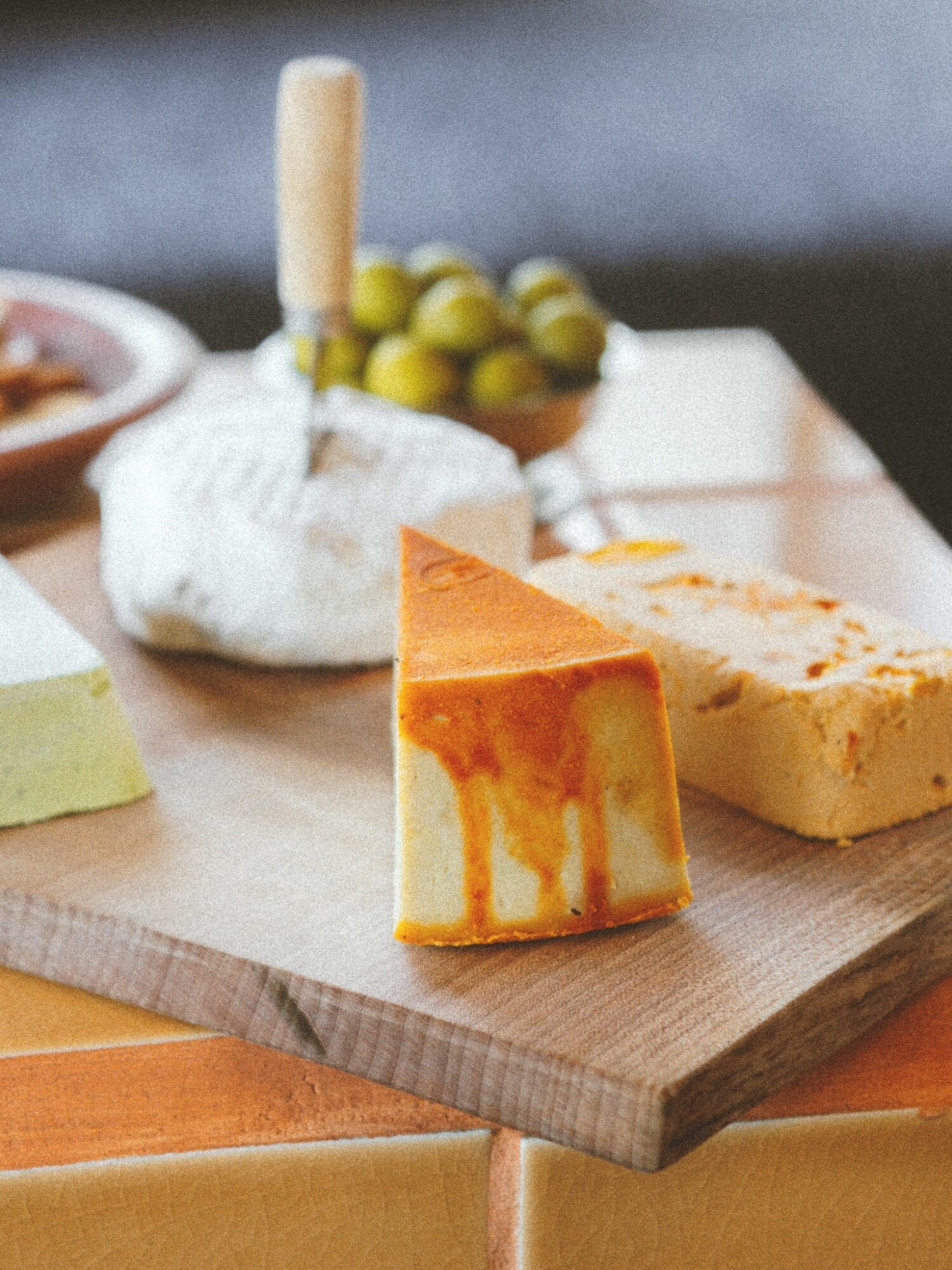 Cheese and other food items on a wooden board, with a blurred background of a bowl of green olives.