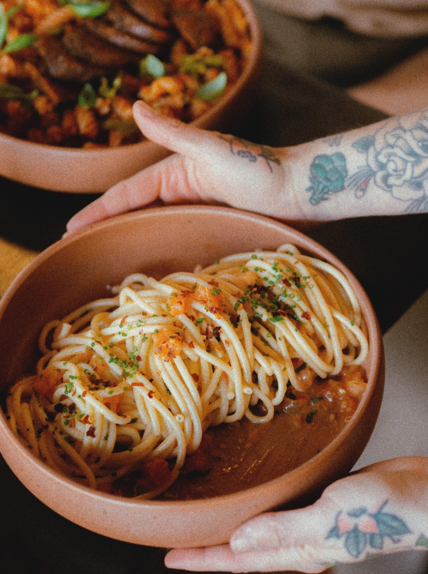 Person with tattooed hands holds a bowl of spaghetti with sauce and herbs, with another dish of food in the background.