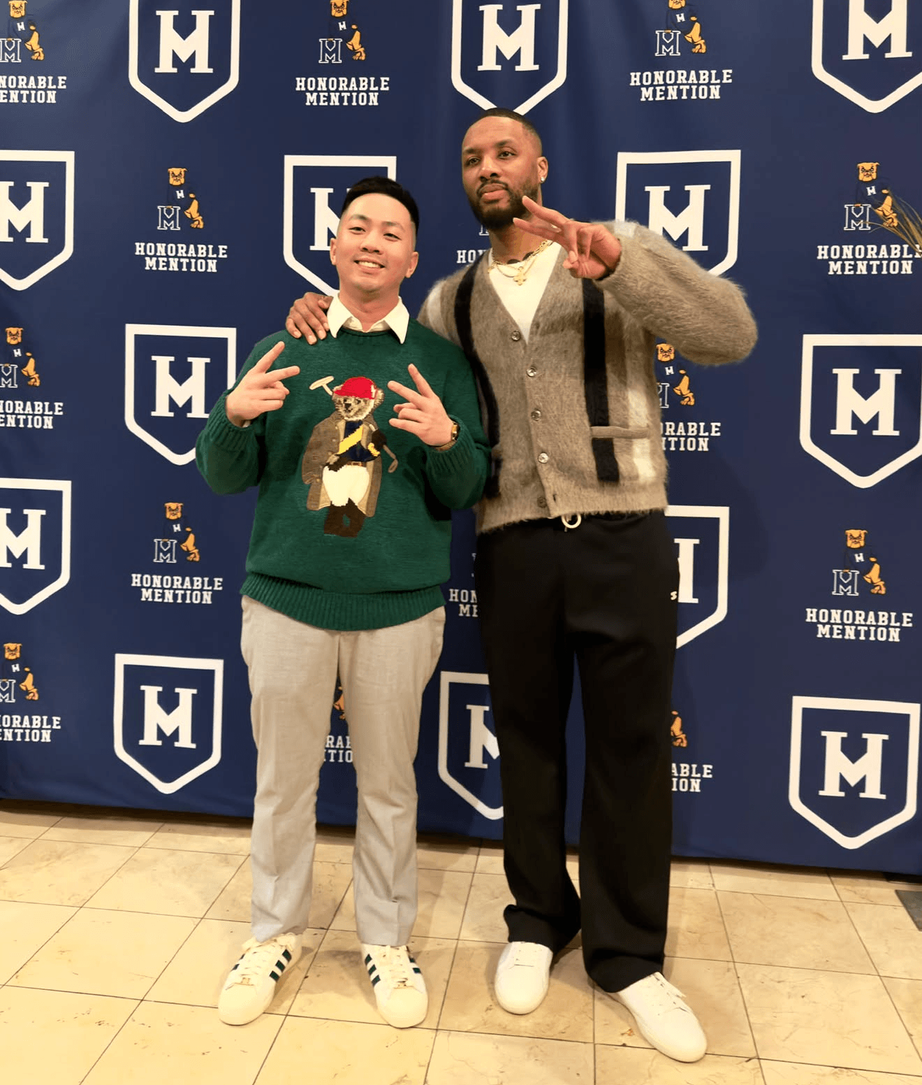Two men standing in front of a blue backdrop with logos, one making a peace sign, the other pointing. Both smiling.