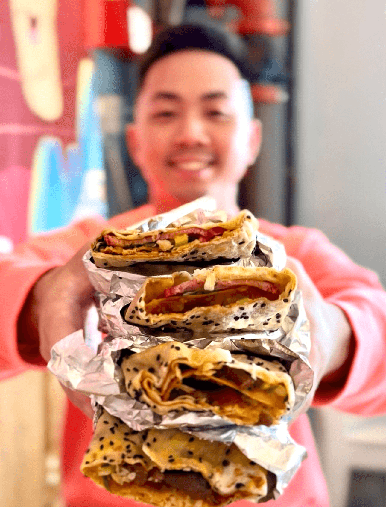 Person holding four stacked sandwiches wrapped in foil, smiling, with colorful background.