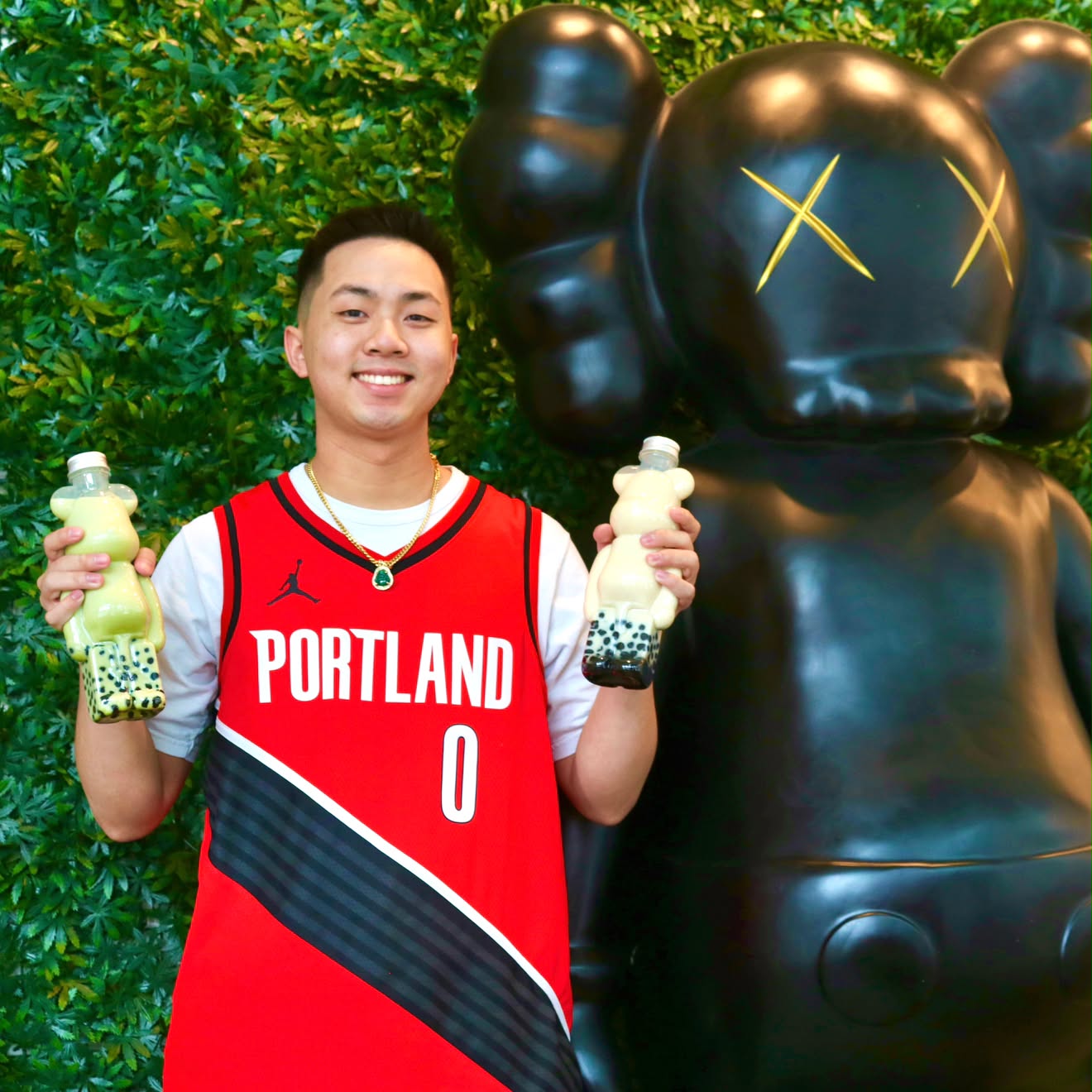 Young man in a red Portland basketball jersey holding bubble tea drinks, standing next to a large black bear statue with yellow X eyes, outdoors with green foliage background.