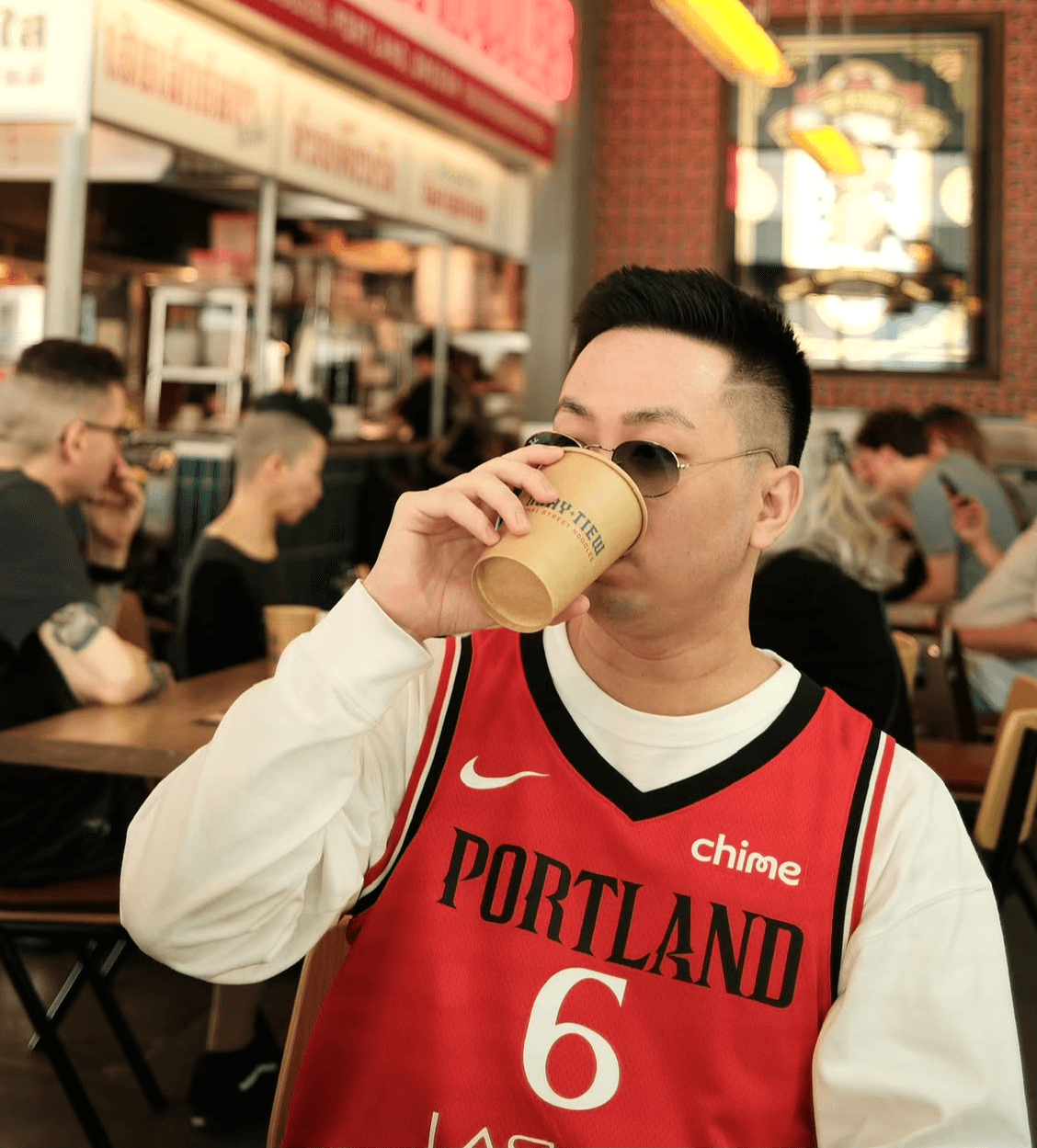 Person wearing a Portland basketball jersey drinking from a paper cup in a busy indoor setting.