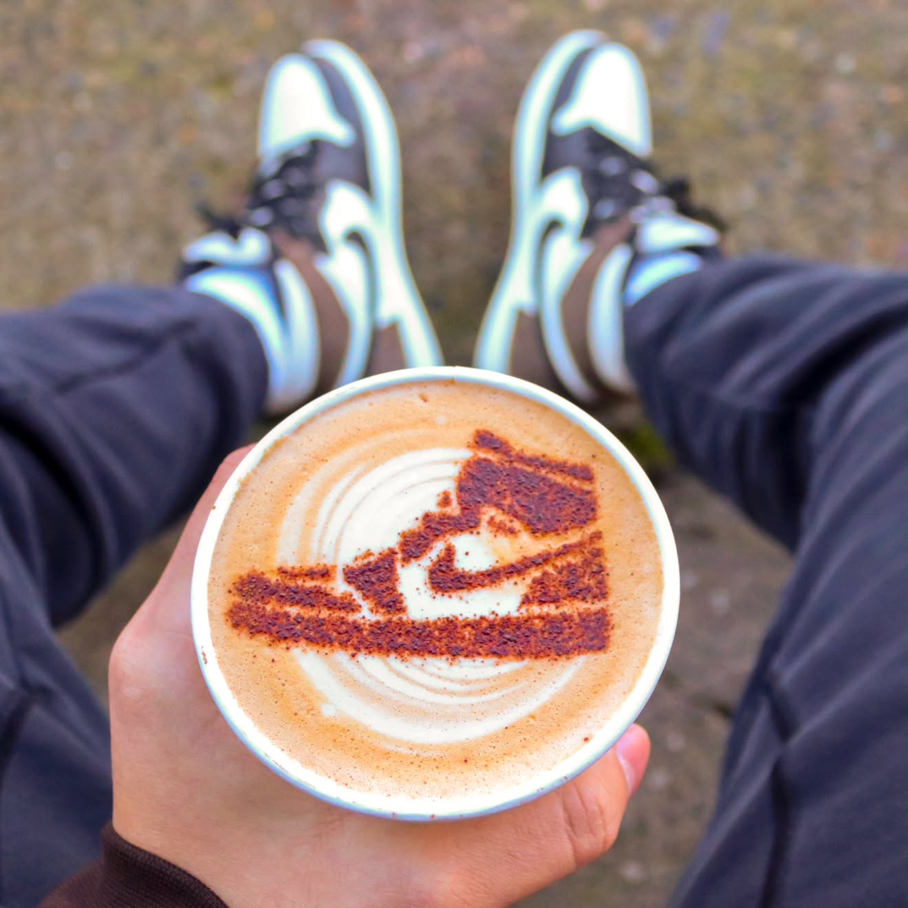 Person sitting outdoors holding a cup of coffee with latte art, wearing sneakers and dark pants.
