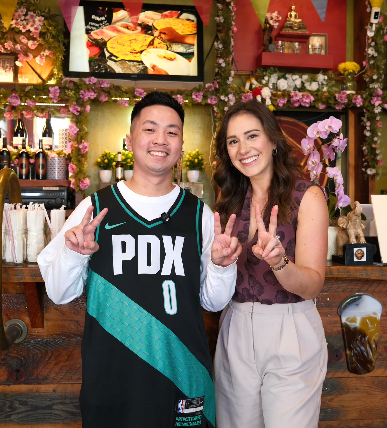 Two people smiling and making peace signs indoors with floral decorations and a bar in the background.