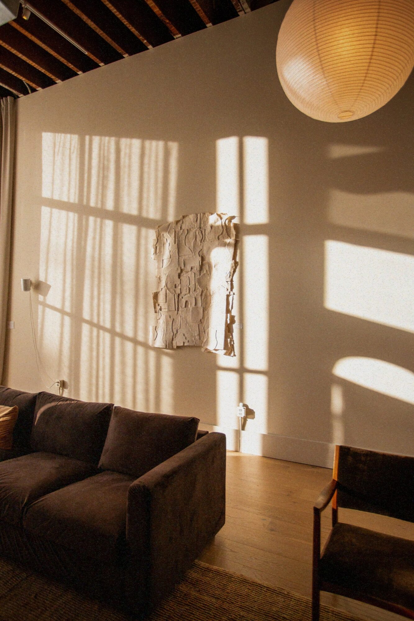 Sunlight creates shadows on a wall with a window and curtain, a hanging paper lantern, and a sofa in a cozy room.