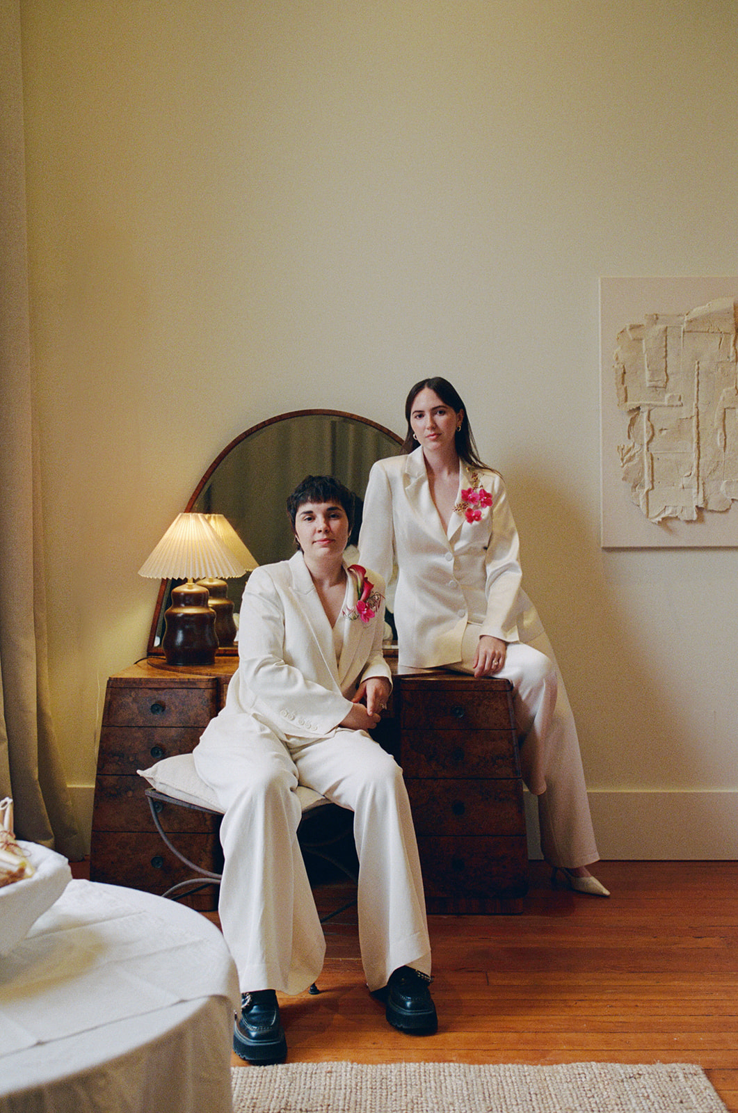 Two women in white suits with pink flowers pose in a room with a mirror, lamp, and artwork on the wall.