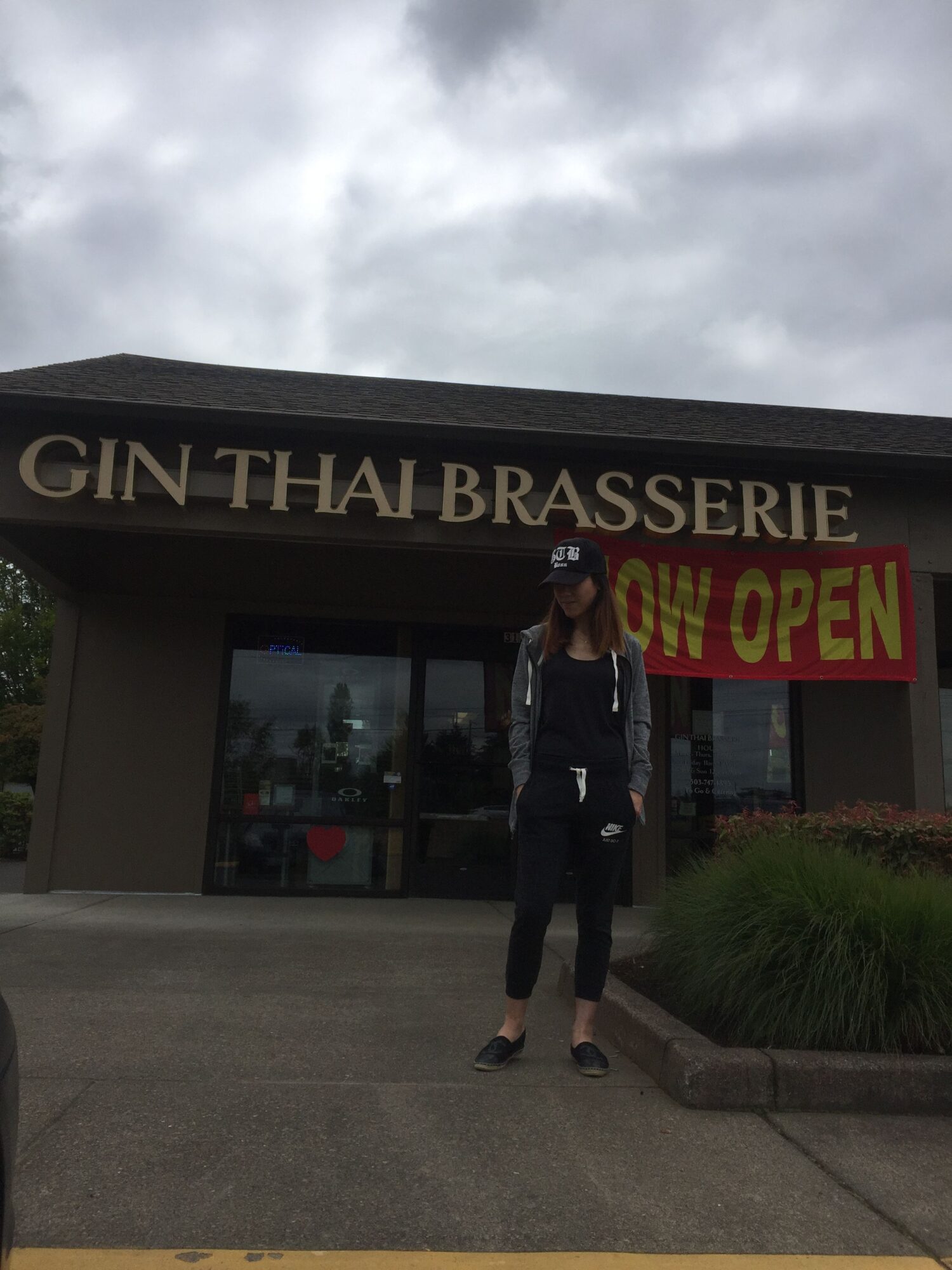 A person stands outside a restaurant entrance beneath a sign reading “GIN THAI BRASSERIE” and “OPEN.”