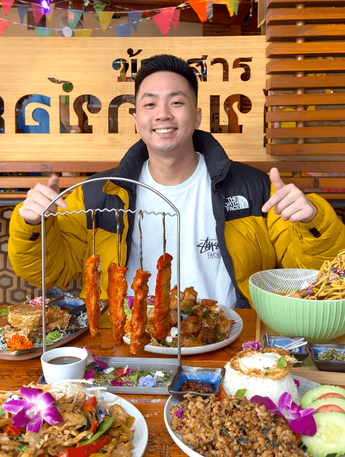 Smiling man in yellow and black jacket sitting at table with food, giving thumbs up, colorful decorations in background.