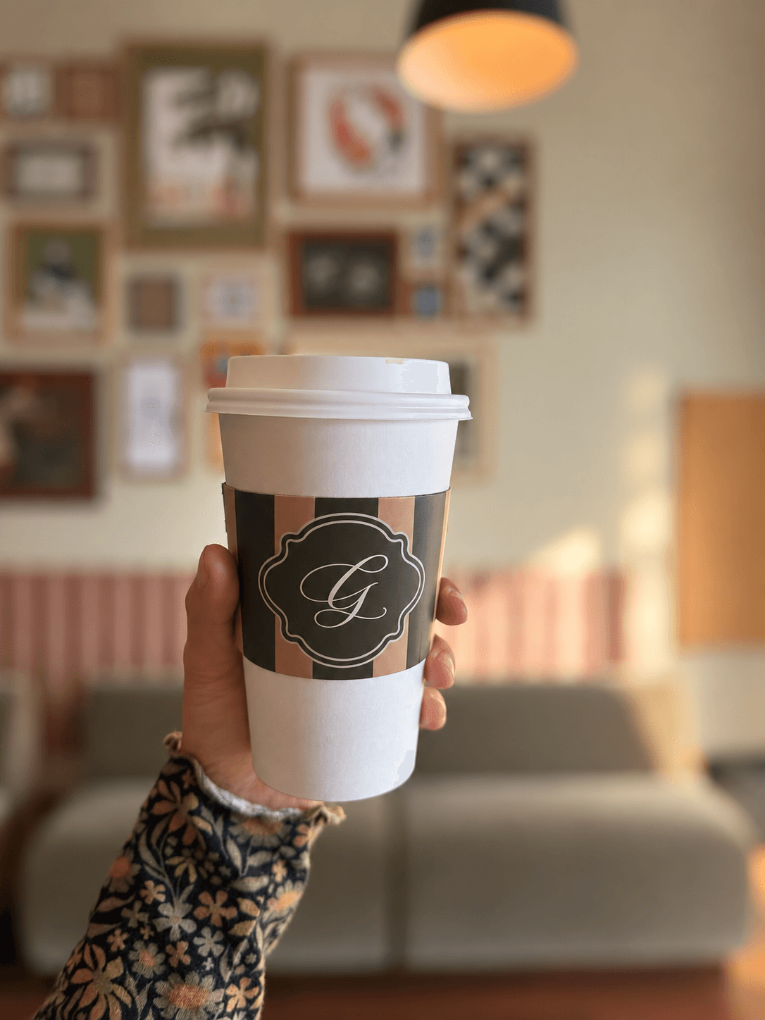 A hand holds a white takeaway cup with a dark decorative logo in a room with framed pictures.