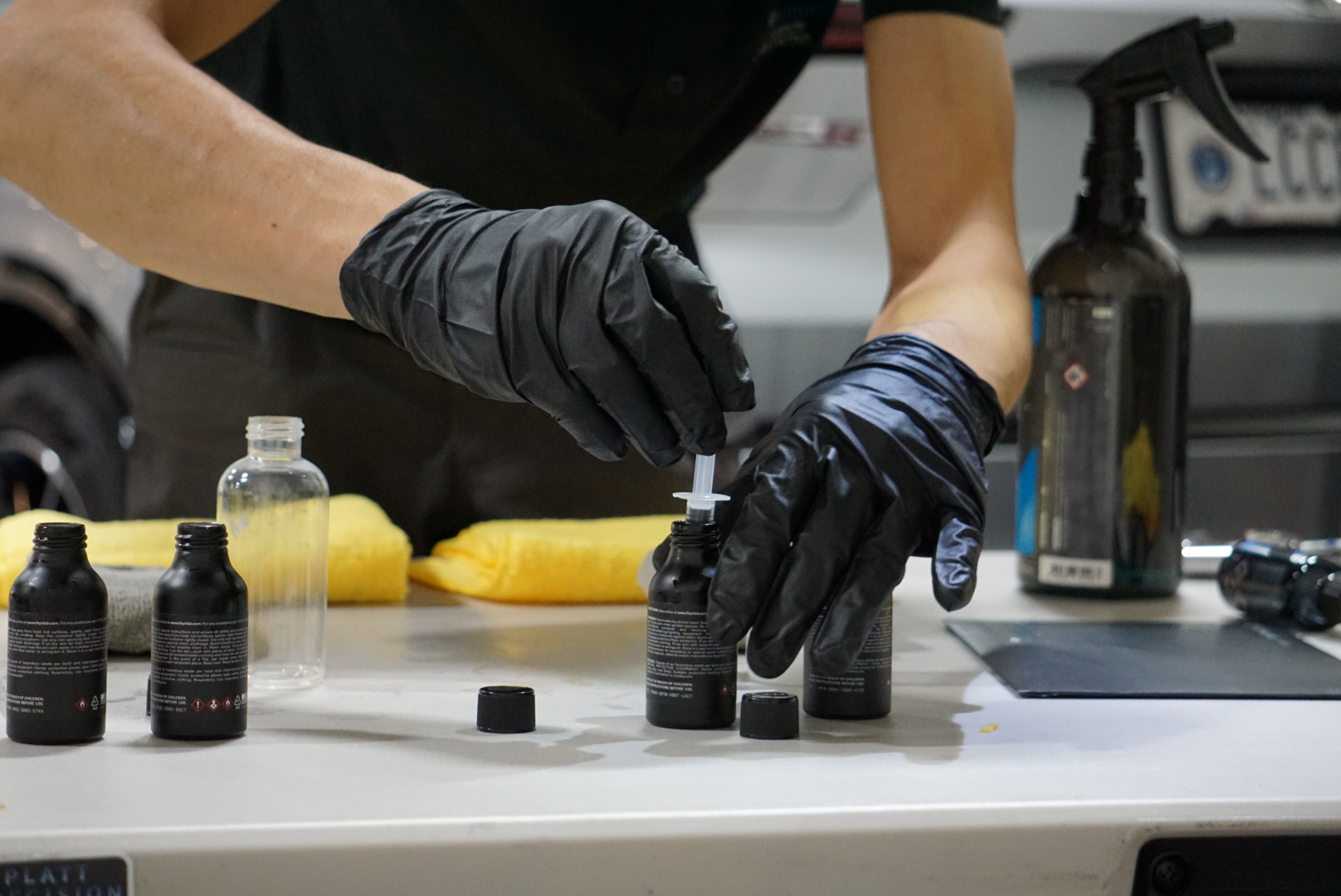 Gloved hands apply liquid from a small bottle at a workbench with other bottles and tools.