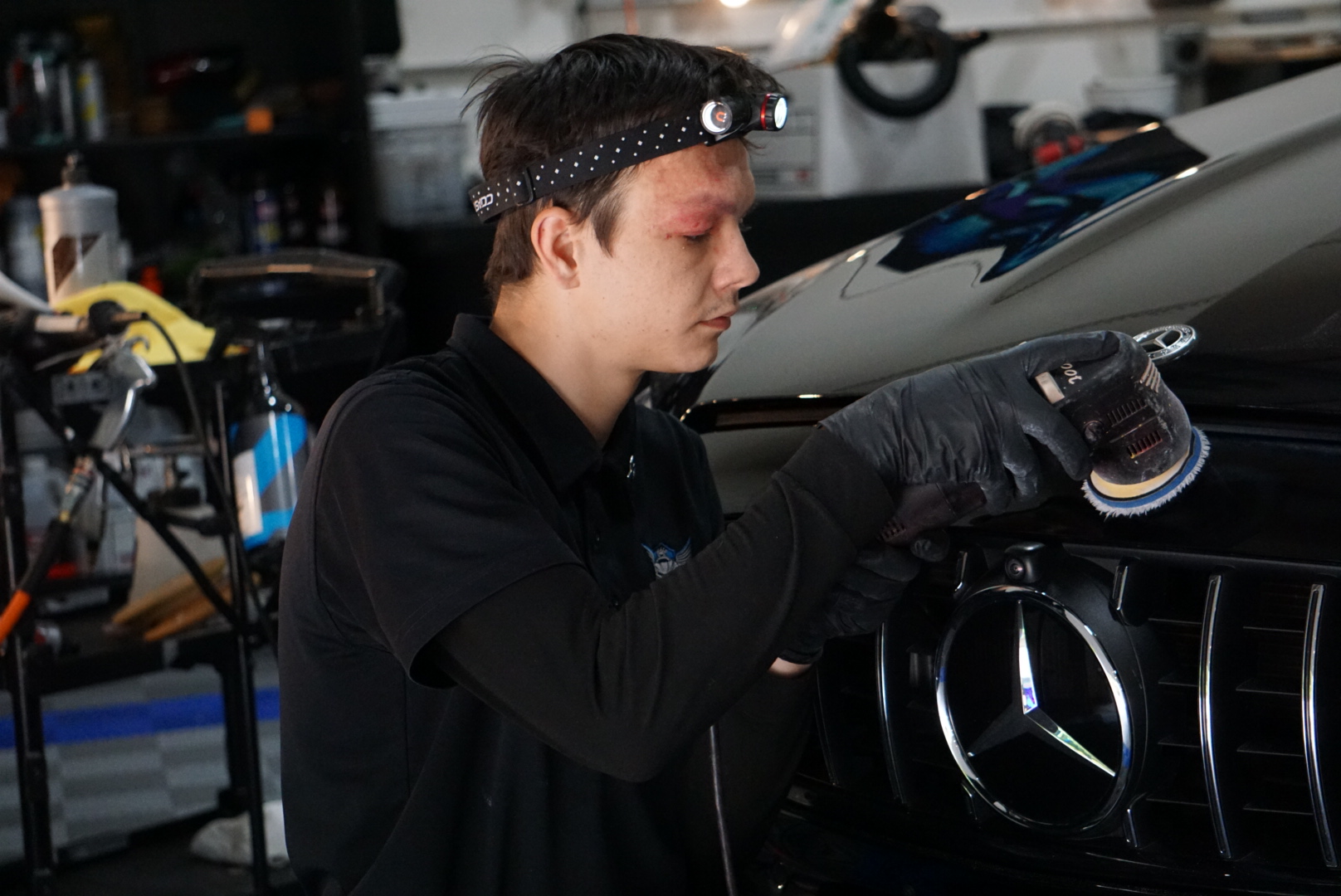 A person wearing a headlamp polishes the front grille of a Mercedes-Benz in a workshop.
