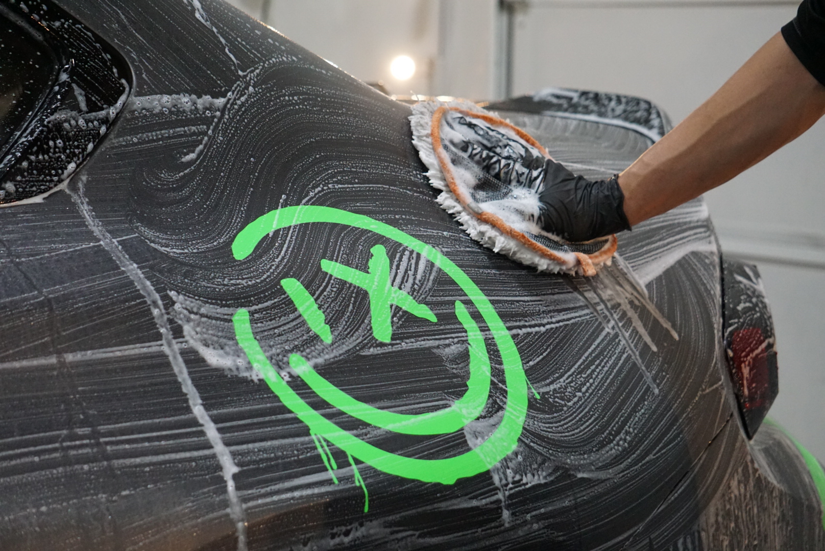 A hand scrubs a soap-covered car marked with a green smiley face.