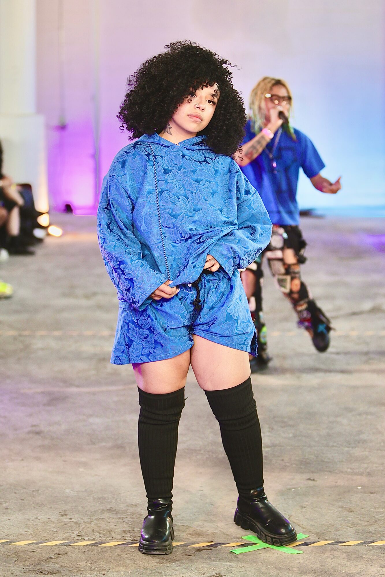 A person in a blue patterned outfit poses on a runway, with another person walking behind.