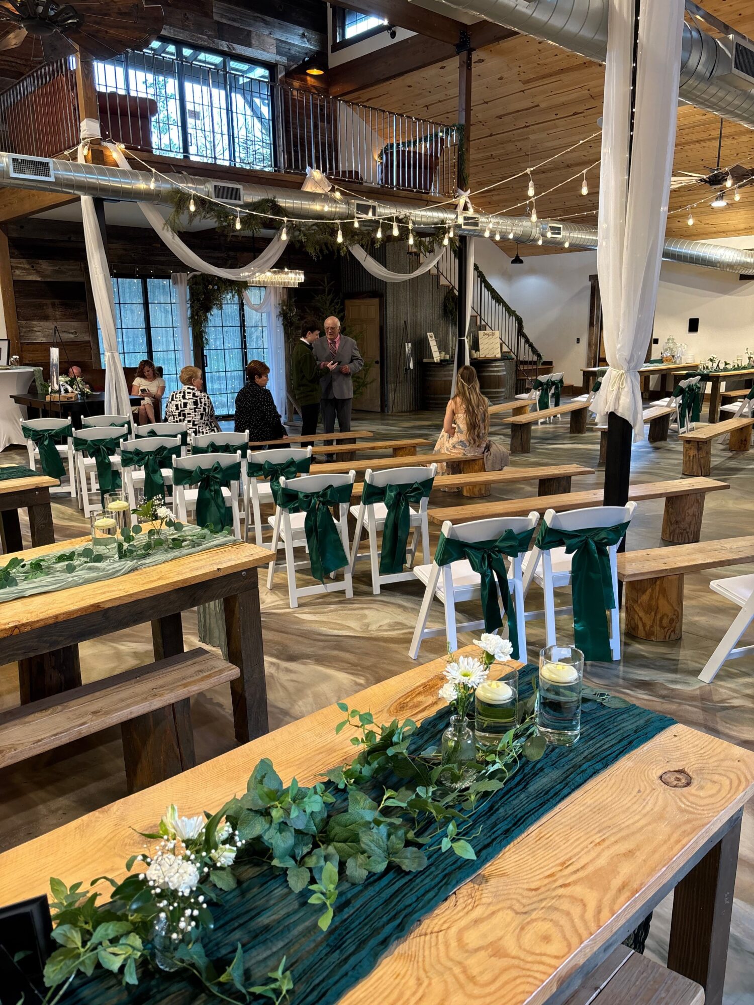 Long wooden dining table decorated with greenery and white flowers, surrounded by chairs in a rustic interior.