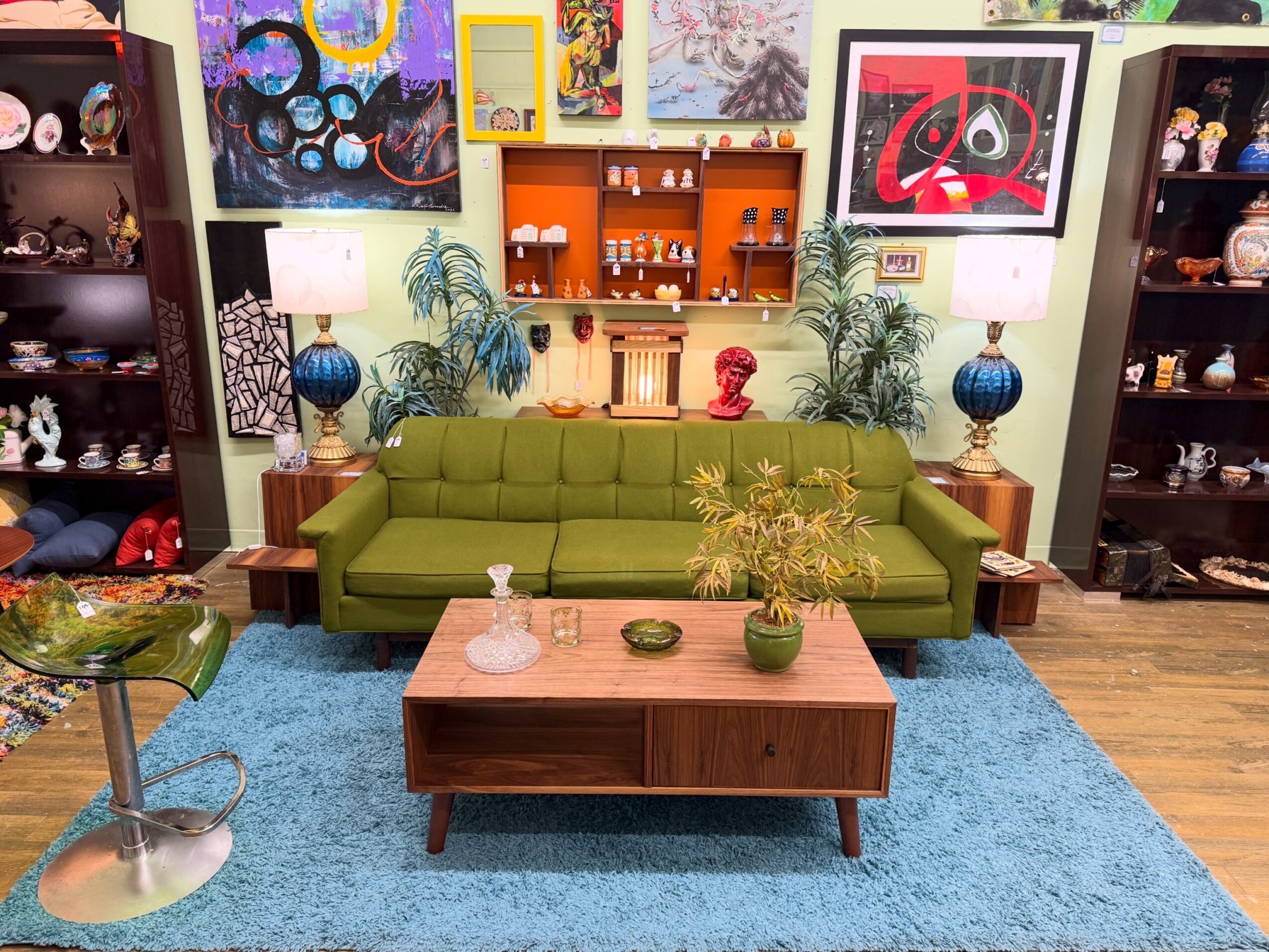 A green sofa and wooden coffee table sit on a blue rug in a room filled with artwork, plants, and display shelves.