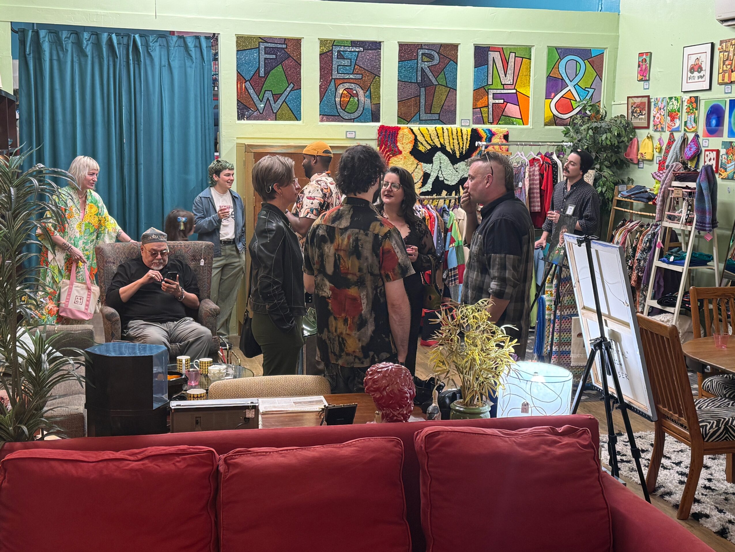 People gather in an art studio filled with colorful paintings, easels, plants, and a red couch.