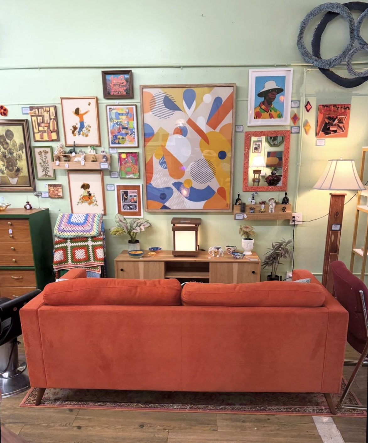 Living room with a red sofa facing a wall of colorful framed artwork, wooden furniture, and a floor lamp.