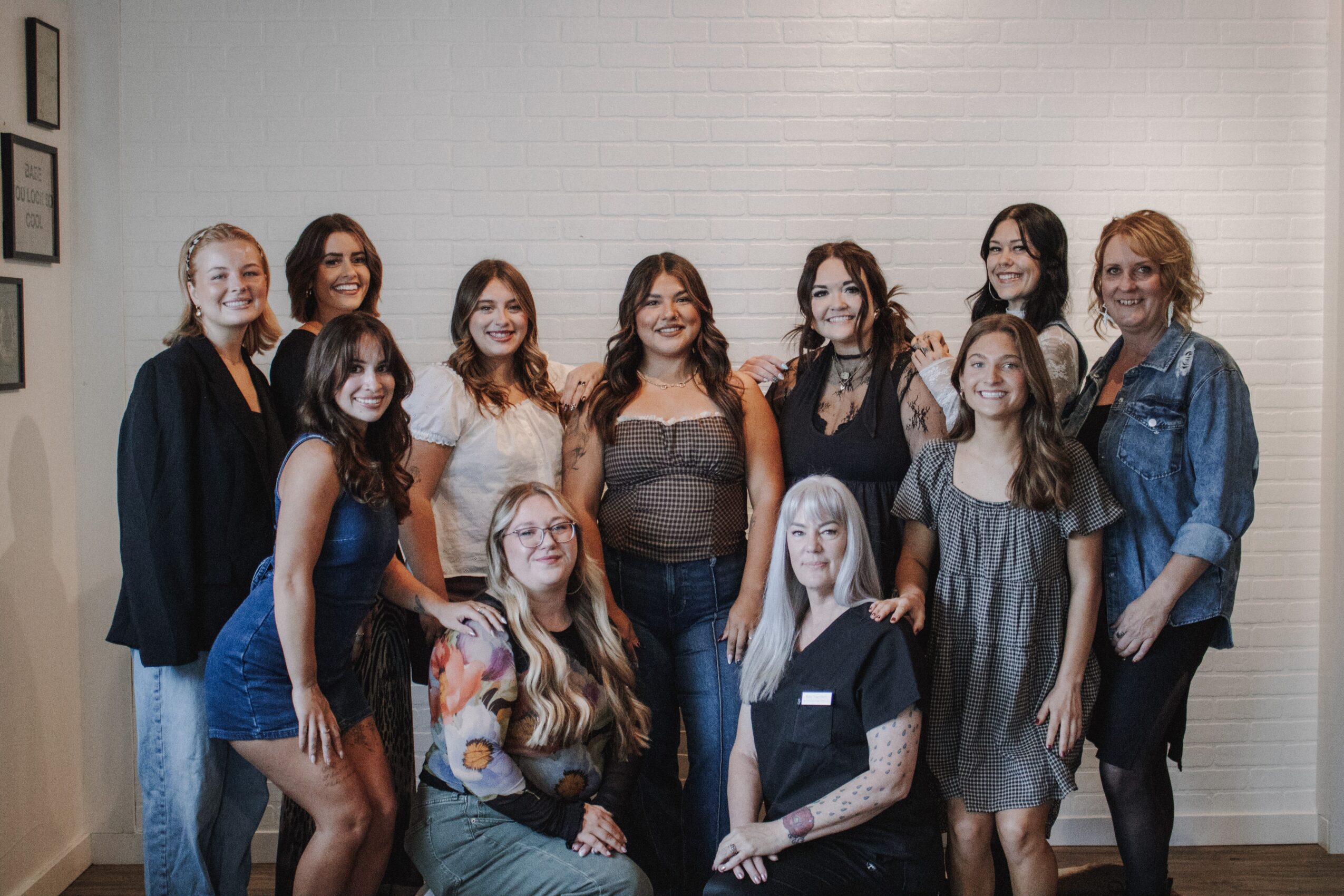 A group of women pose together in two rows in front of a light brick wall.