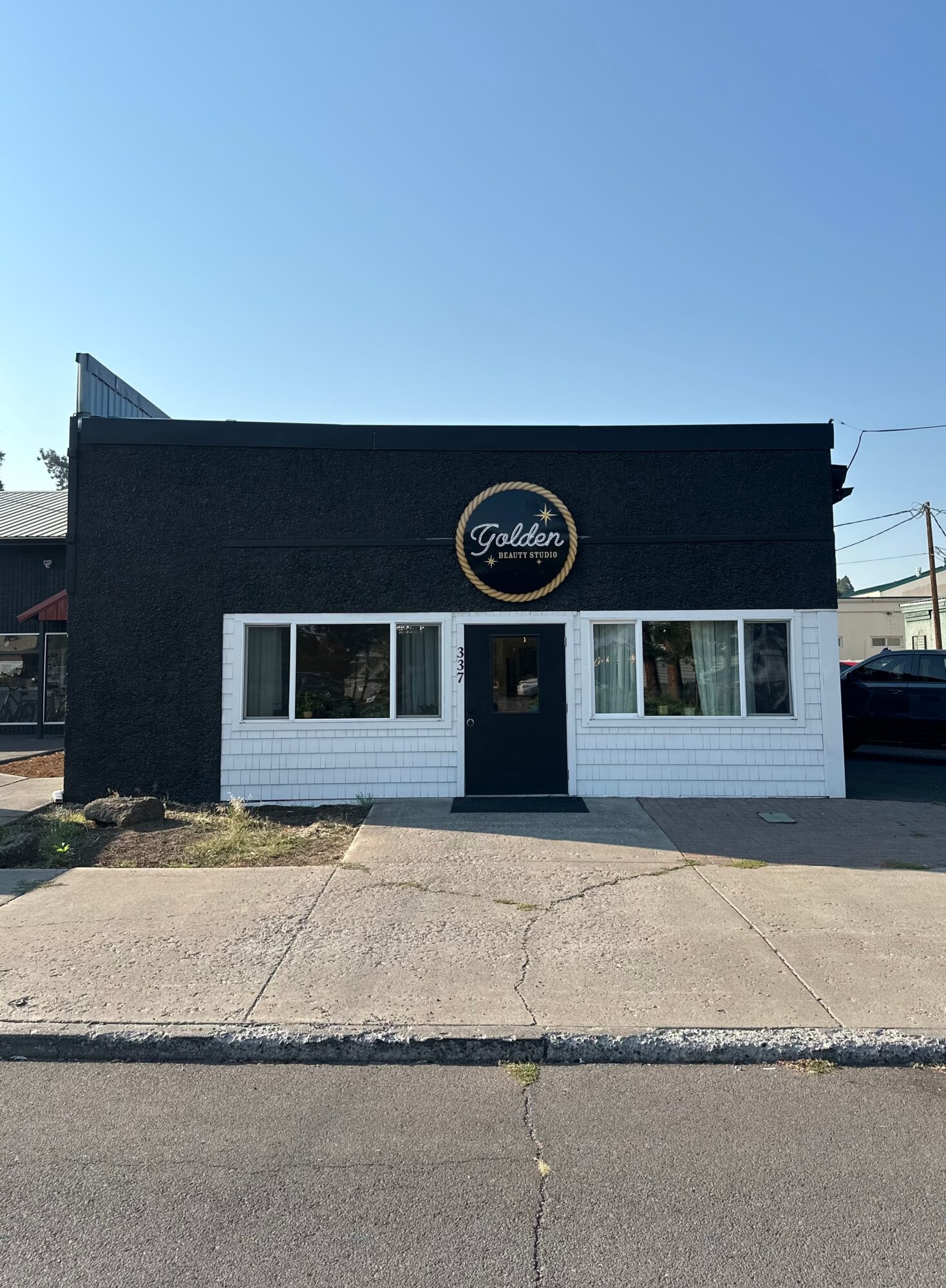 Small black storefront with white windows and a circular sign, beside a concrete sidewalk under a clear blue sky.