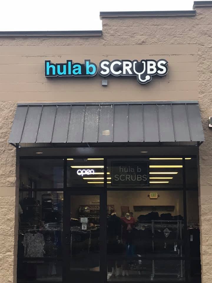 Storefront with a sign reading “hula b SCRUBS” above a dark awning and glass entrance.