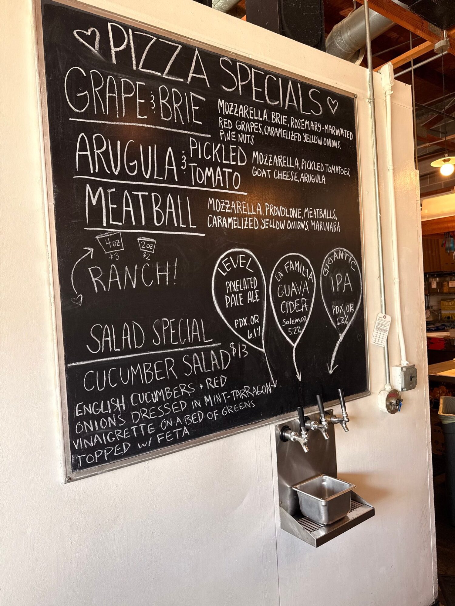 Chalkboard menu listing pizza, grape brie, ravioli, meatball, ranch, and salad beside a metal dispenser.