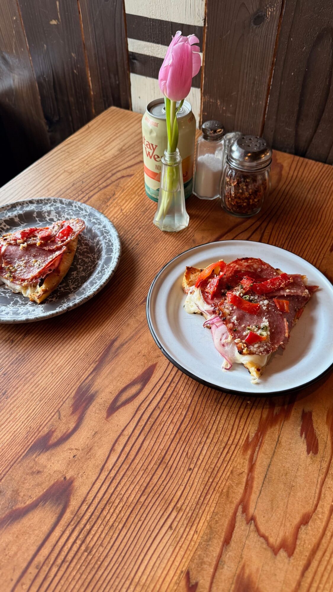 Two plates of food on a wooden table, with a pink flower in a vase and small jars nearby.
