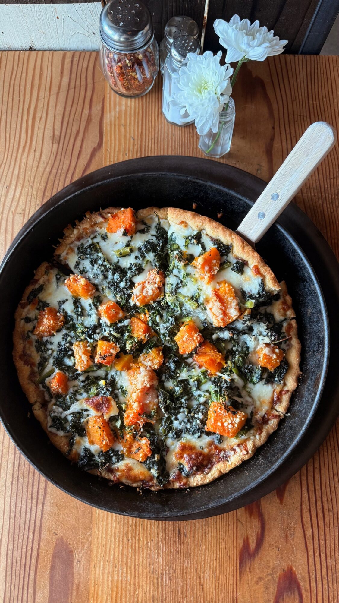 A vegetable-topped pizza in a black skillet on a wooden table, with a metal server and small jars nearby.
