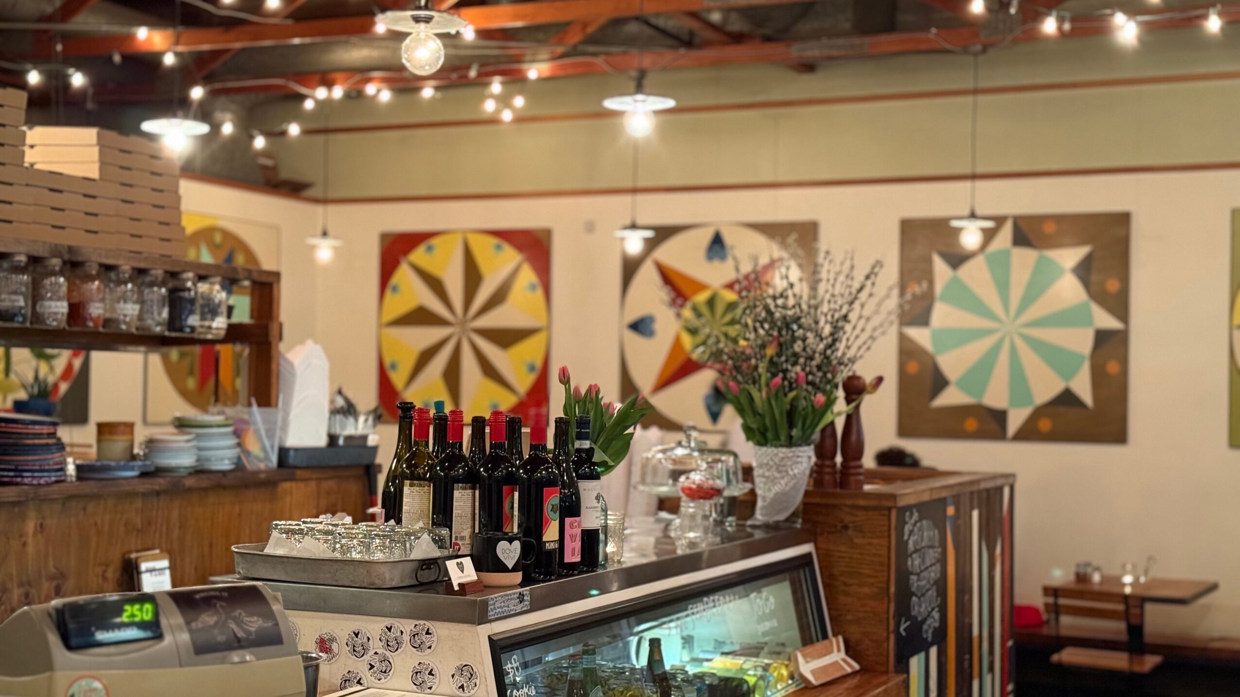A restaurant counter with wine bottles, glass display cases, flowers, and colorful geometric wall art.