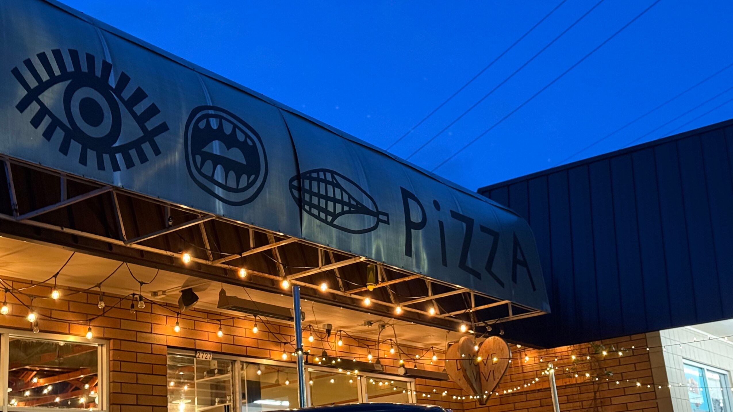 Street-level view of a restaurant facade with an eye graphic, a pizza sign, and illuminated windows.
