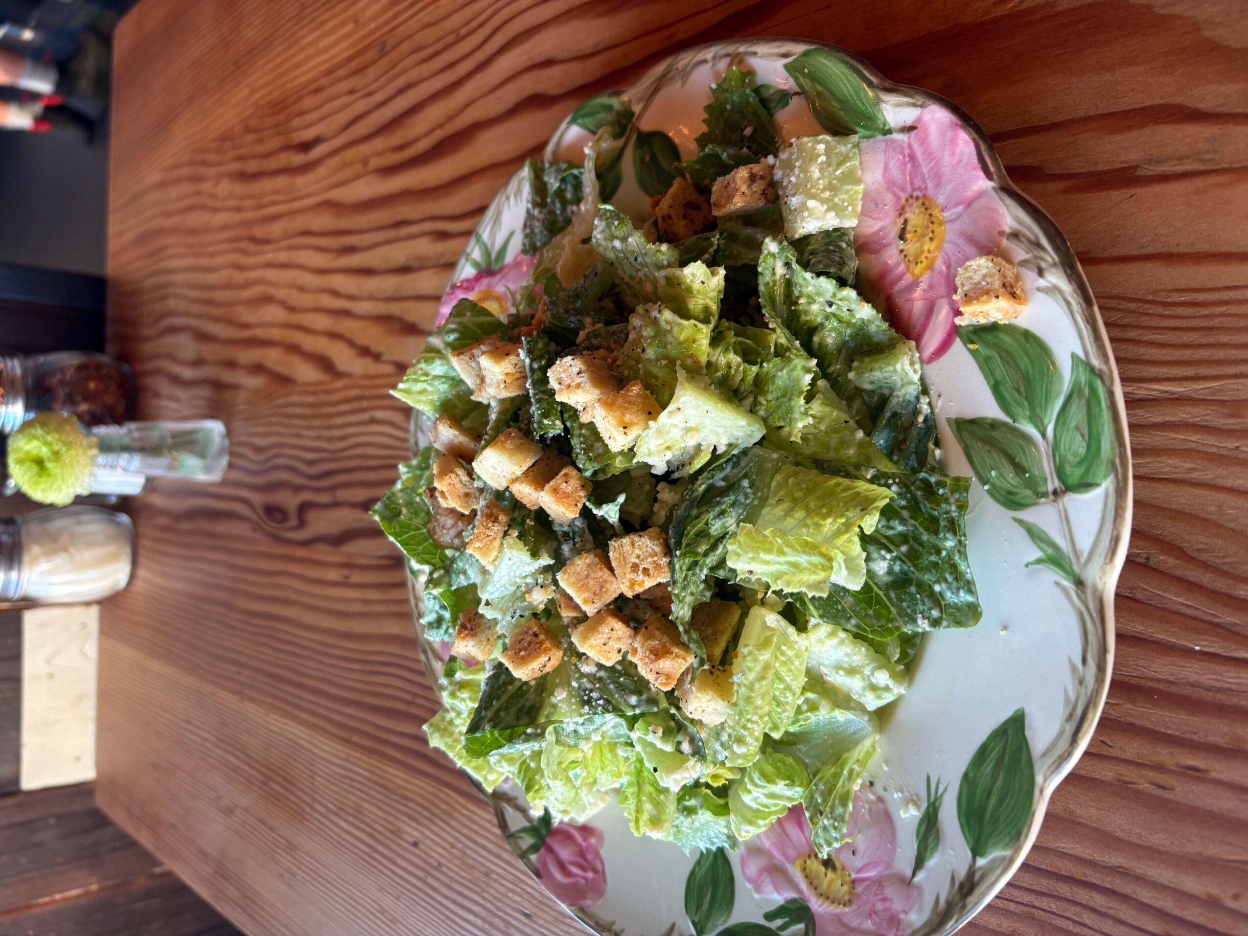 A bowl of leafy salad with croutons and dressing on a floral plate atop a wooden table.