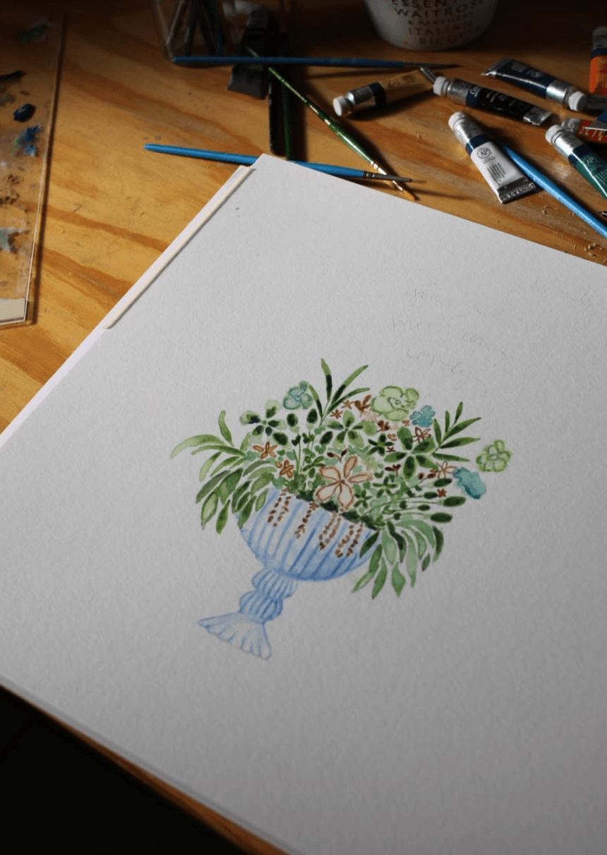 A watercolor floral arrangement in a blue vase on paper beside art supplies on a wooden desk.