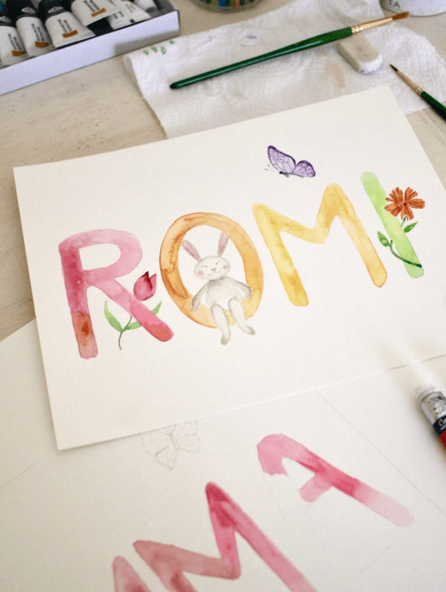 Watercolor painting of the word “ROMA” with a rabbit, butterfly, and flower, beside art supplies.