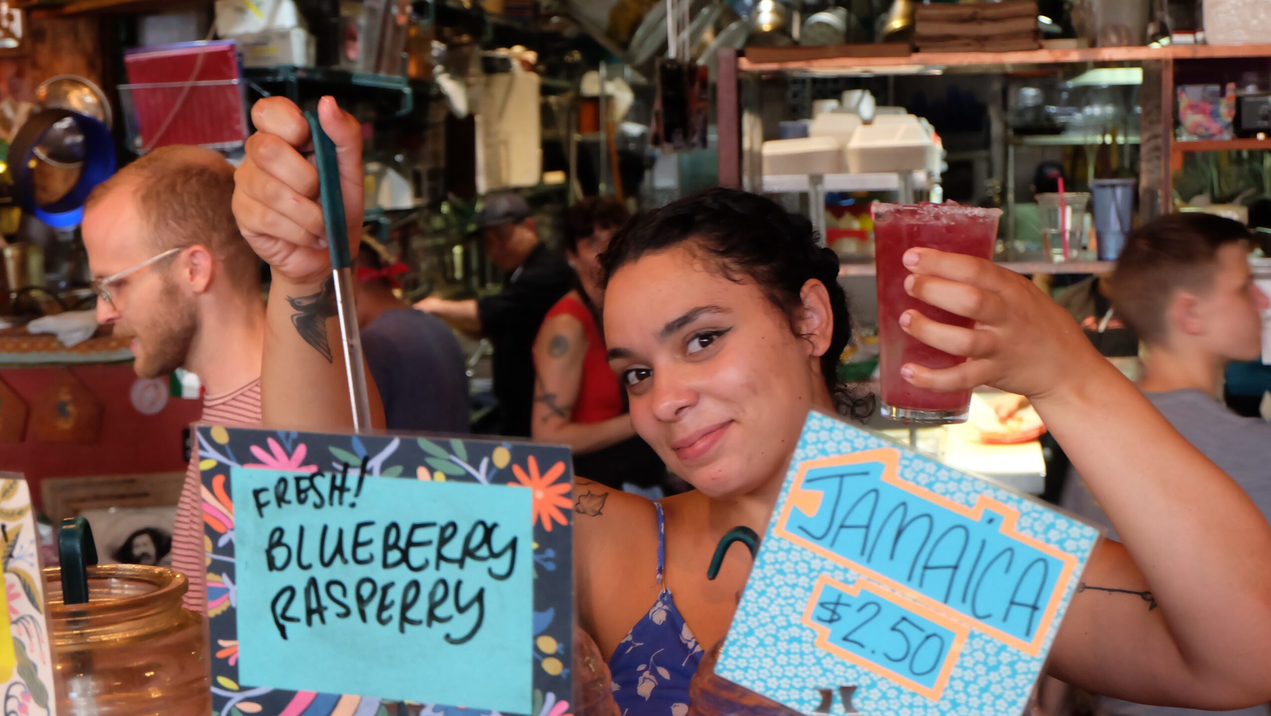 A person holds signs advertising fresh blueberry raspberry and Jamaica drinks in a crowded market.