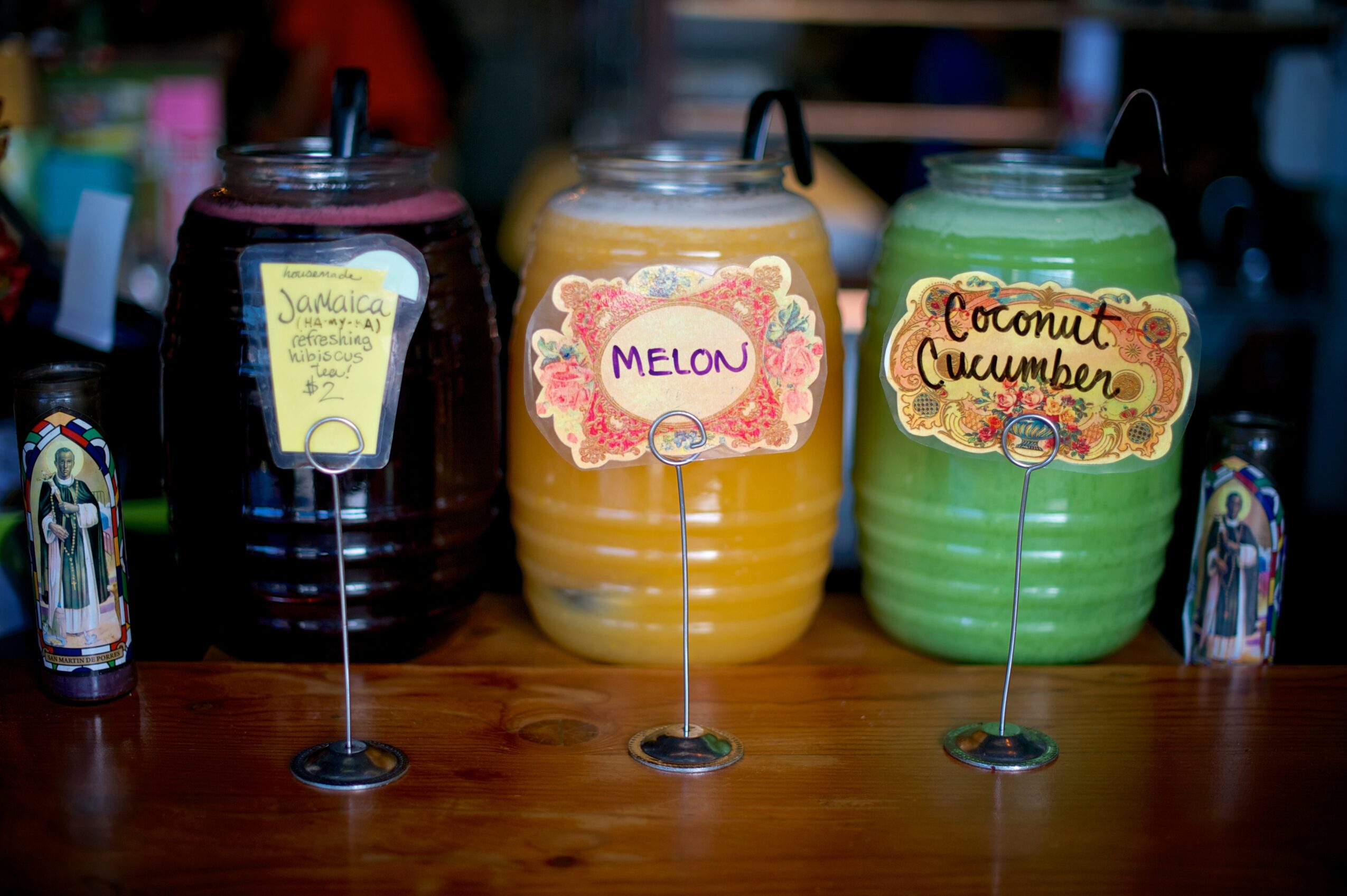 Three large beverage dispensers labeled Jamaican, Melon, and Coconut Cucumber sit on a wooden counter.