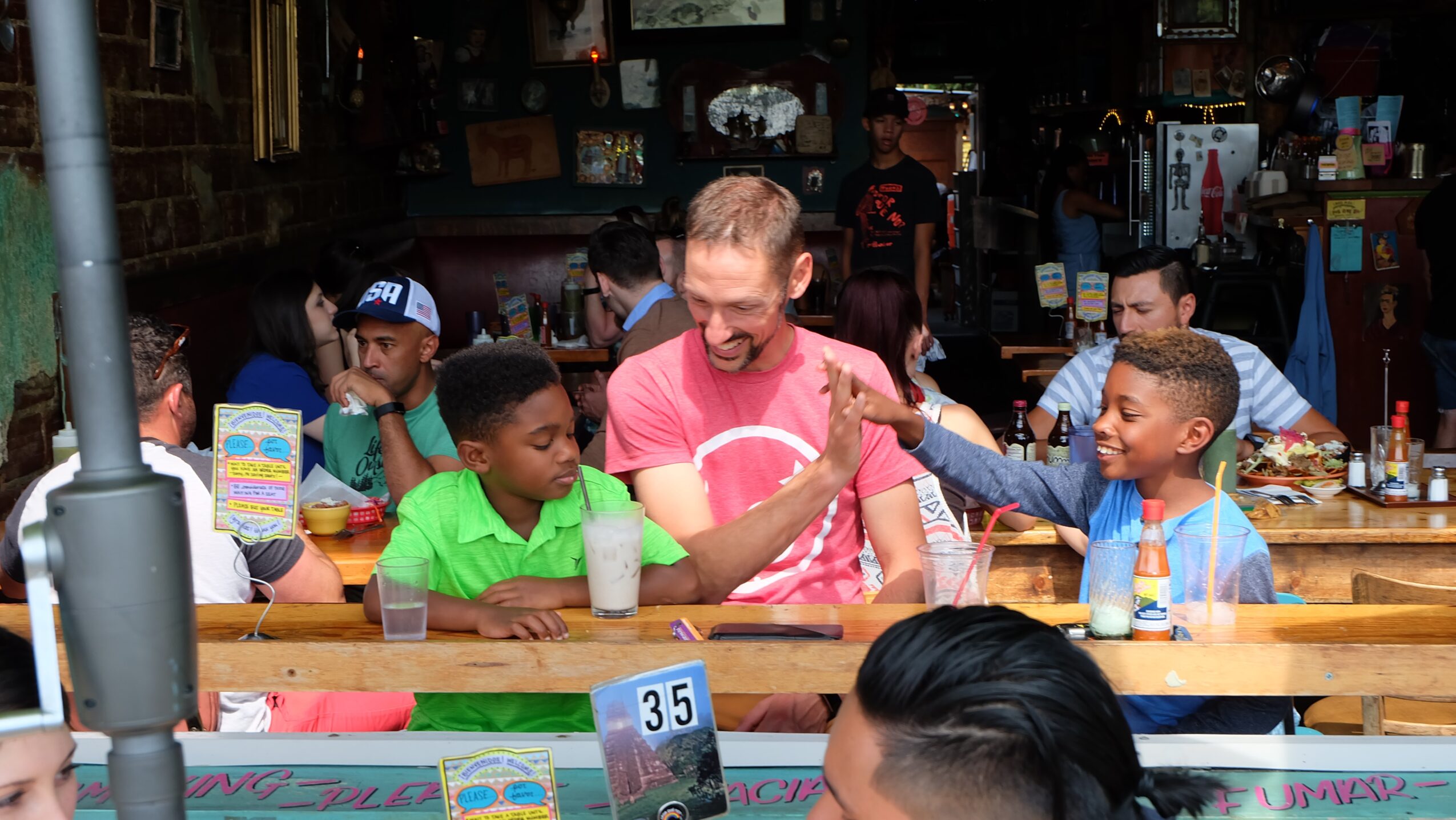A man in a pink shirt sits at a table with several people, including children, in a shop.