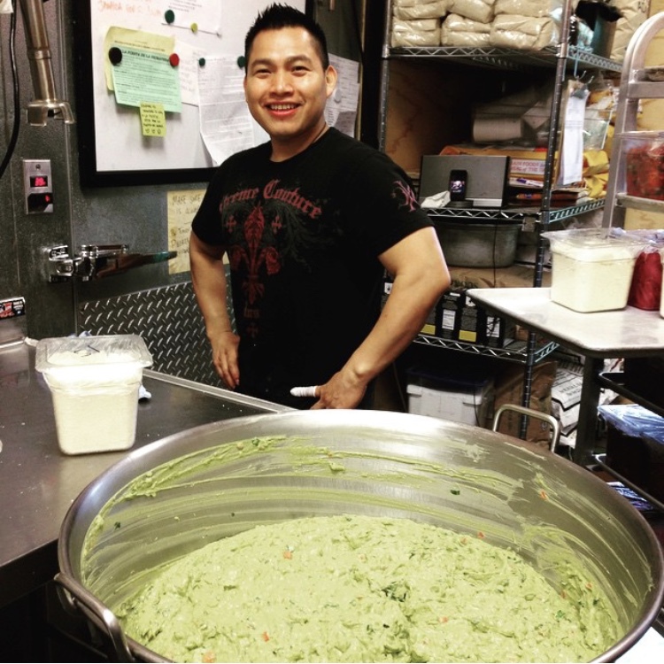 Person standing behind a large metal bowl filled with a pale green mixture in a commercial kitchen.
