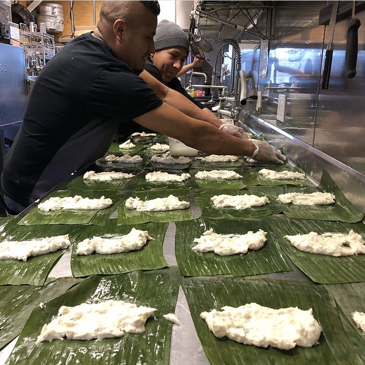 Two people prepare portions of food on banana leaves along a stainless-steel worktable.