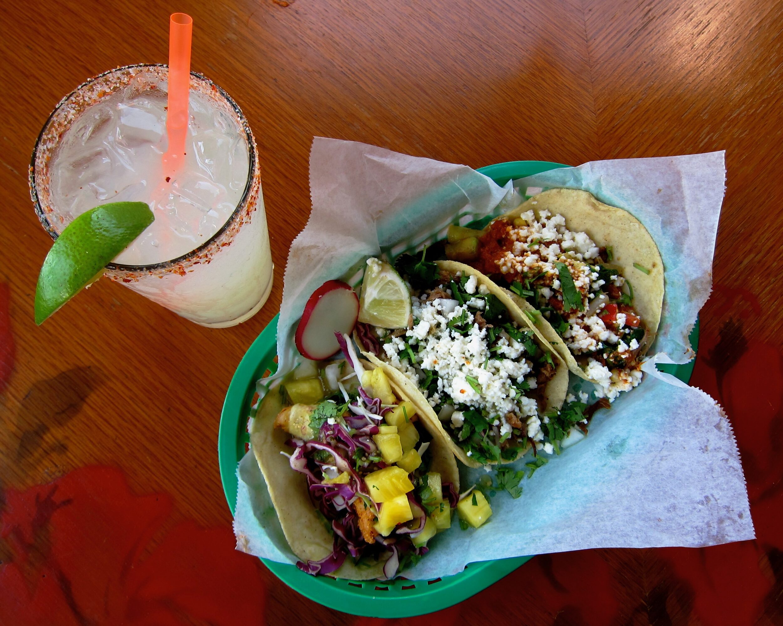 A basket with two tacos topped with vegetables and crumbled cheese, beside a drink with a lime wedge and orange straw.