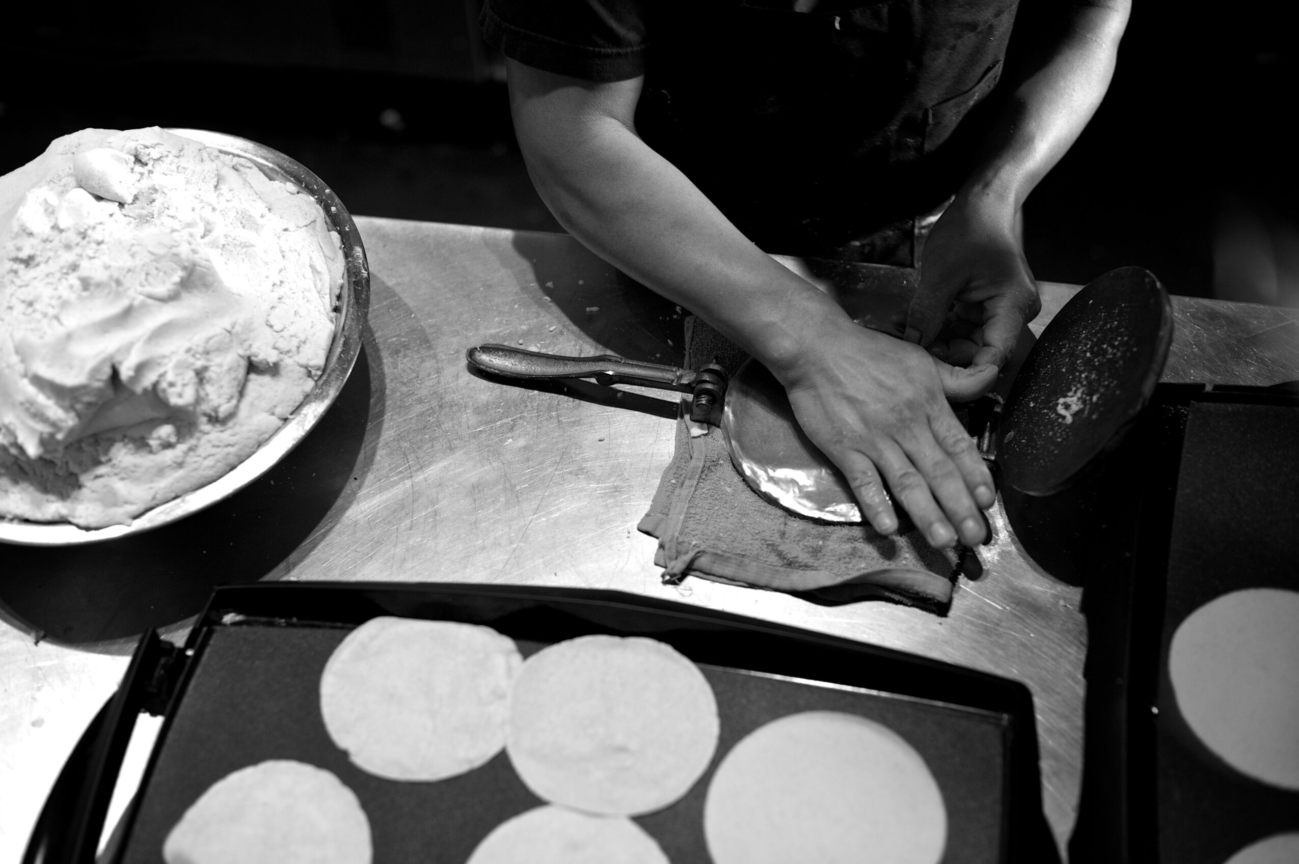 A person shapes dough beside a bowl of flour and a tray of round flatbreads.