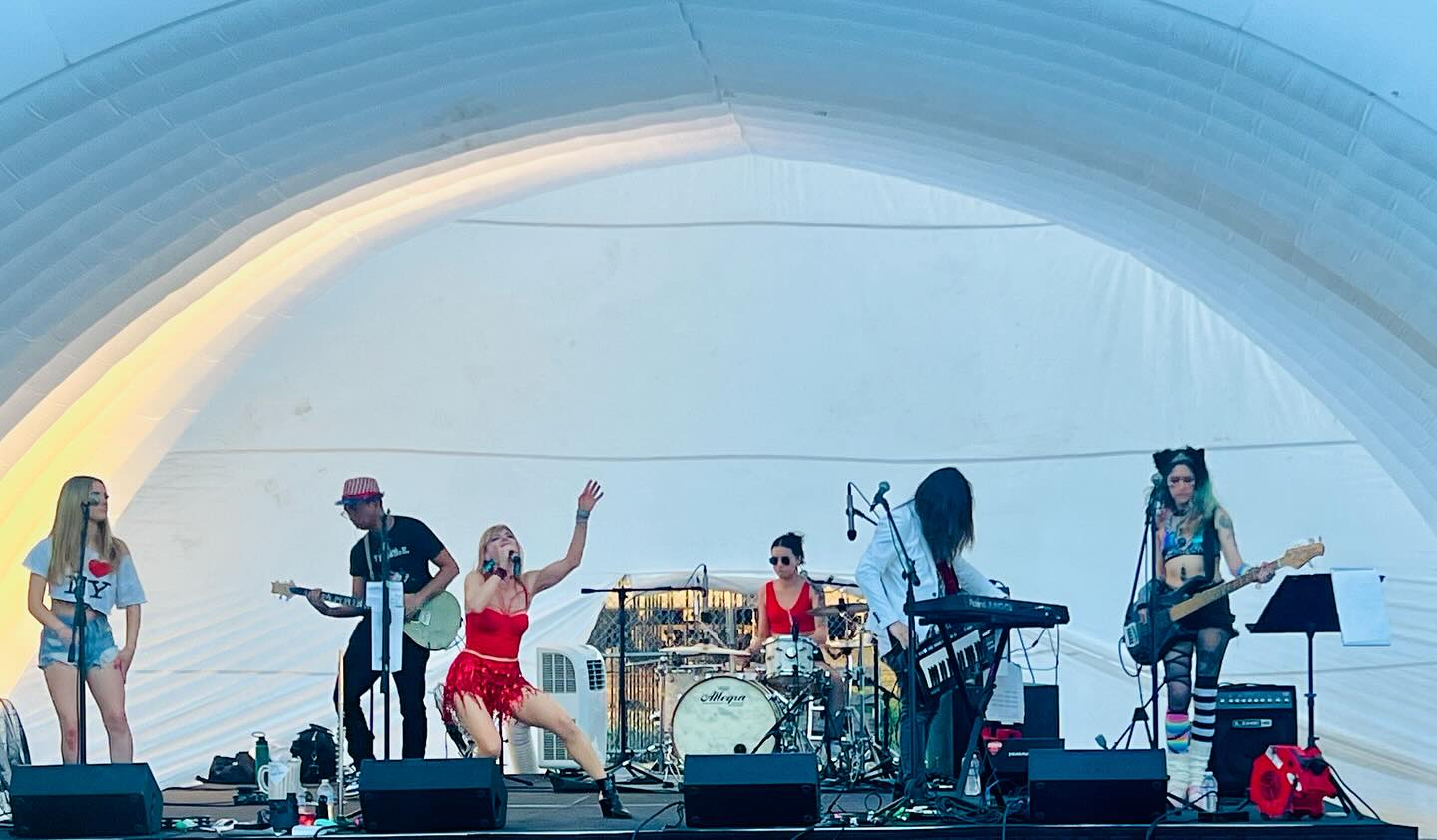 A band performs on an outdoor stage beneath a curved canopy, with musicians, instruments, and stage equipment.