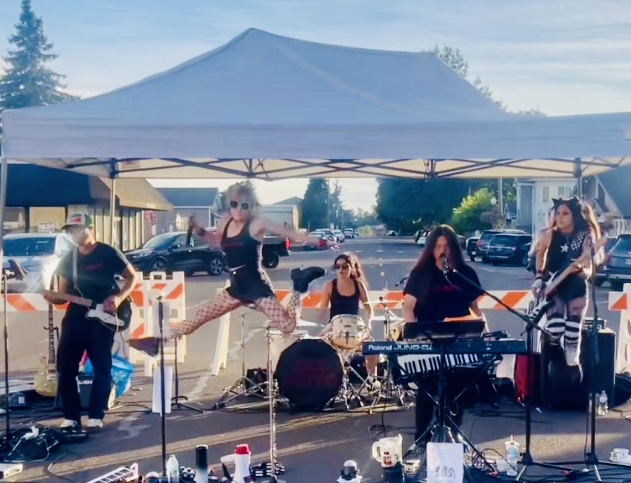 A band performs beneath a canopy outdoors, with a singer, musicians, instruments, and audio equipment.