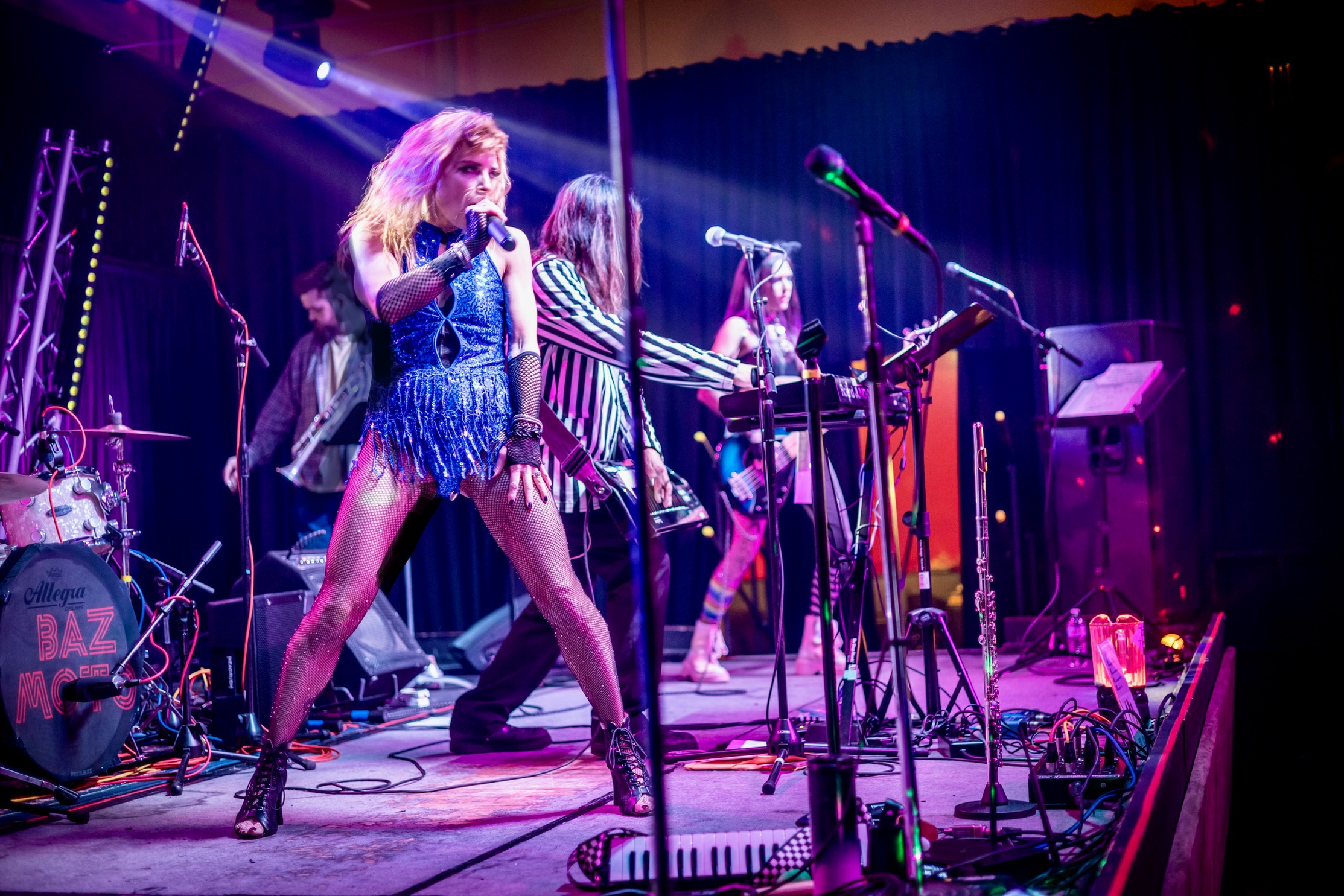 A singer performs onstage with musicians, microphones, instruments, and colorful stage lighting.